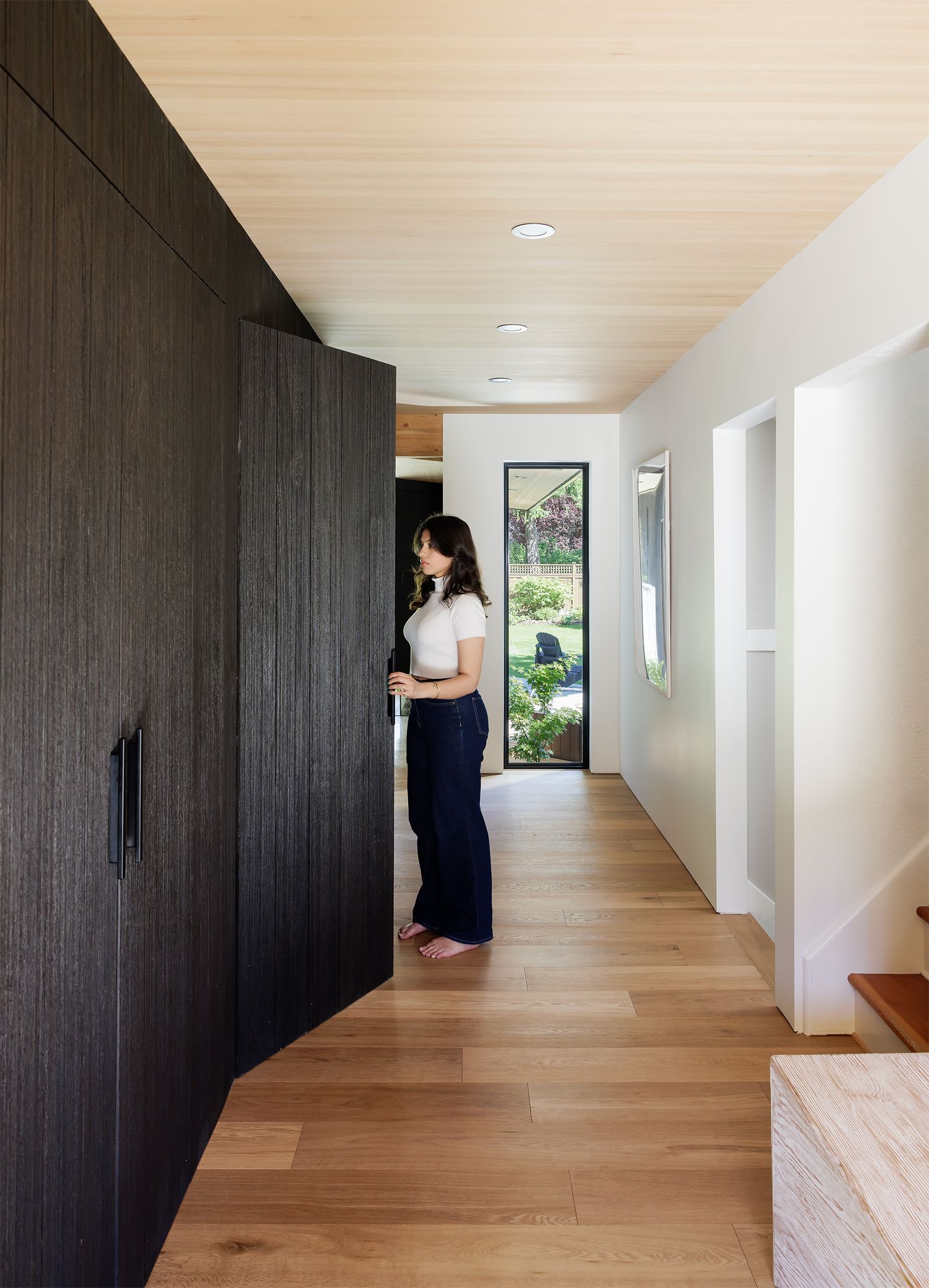 Modern hallway with dark wood panel storage wall, light oak floors, wood ceiling, and garden view through glass door.