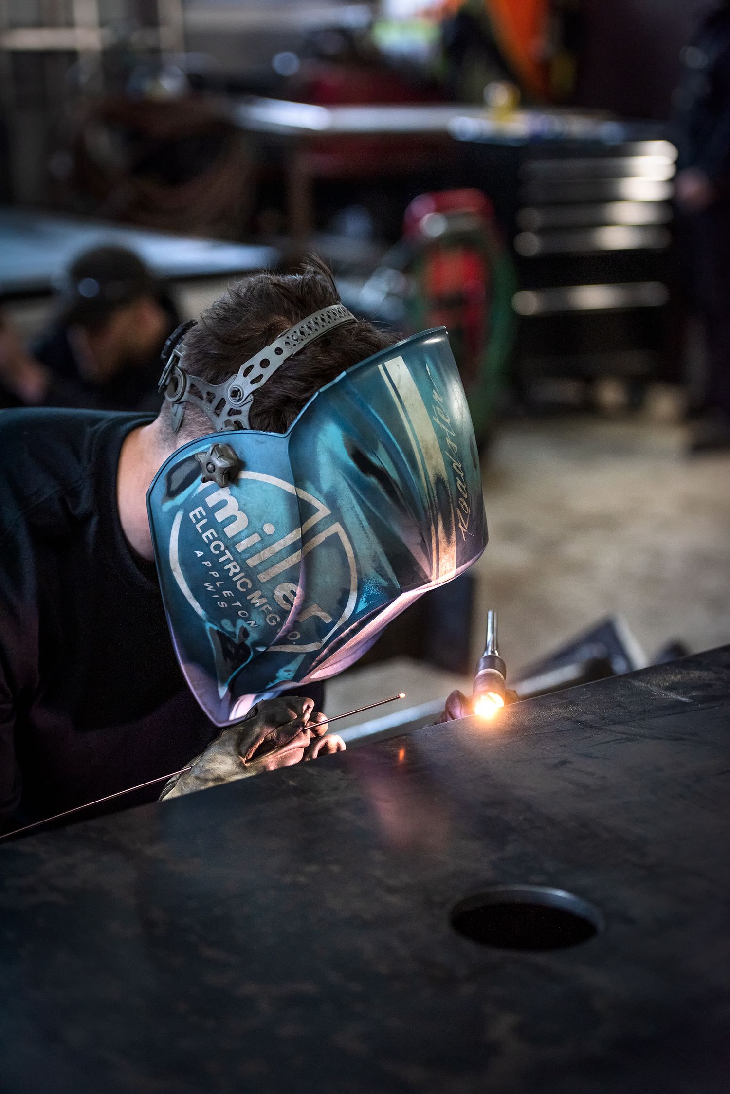 Close-up of welder wearing Miller Electric helmet welding metal in a dark industrial workshop.