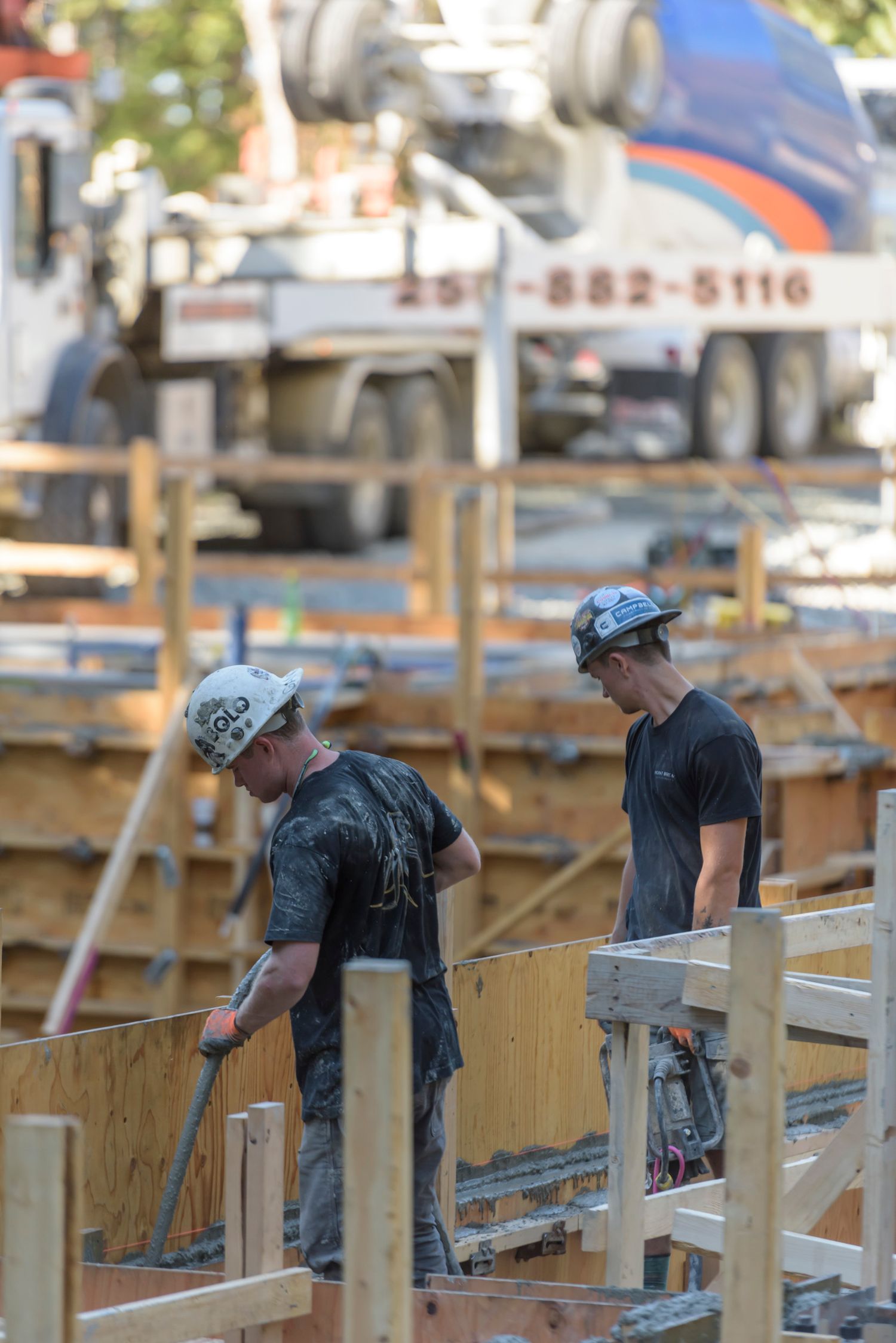 Two construction workers in hard hats work with tools at a concrete forming site with a pump truck behind.