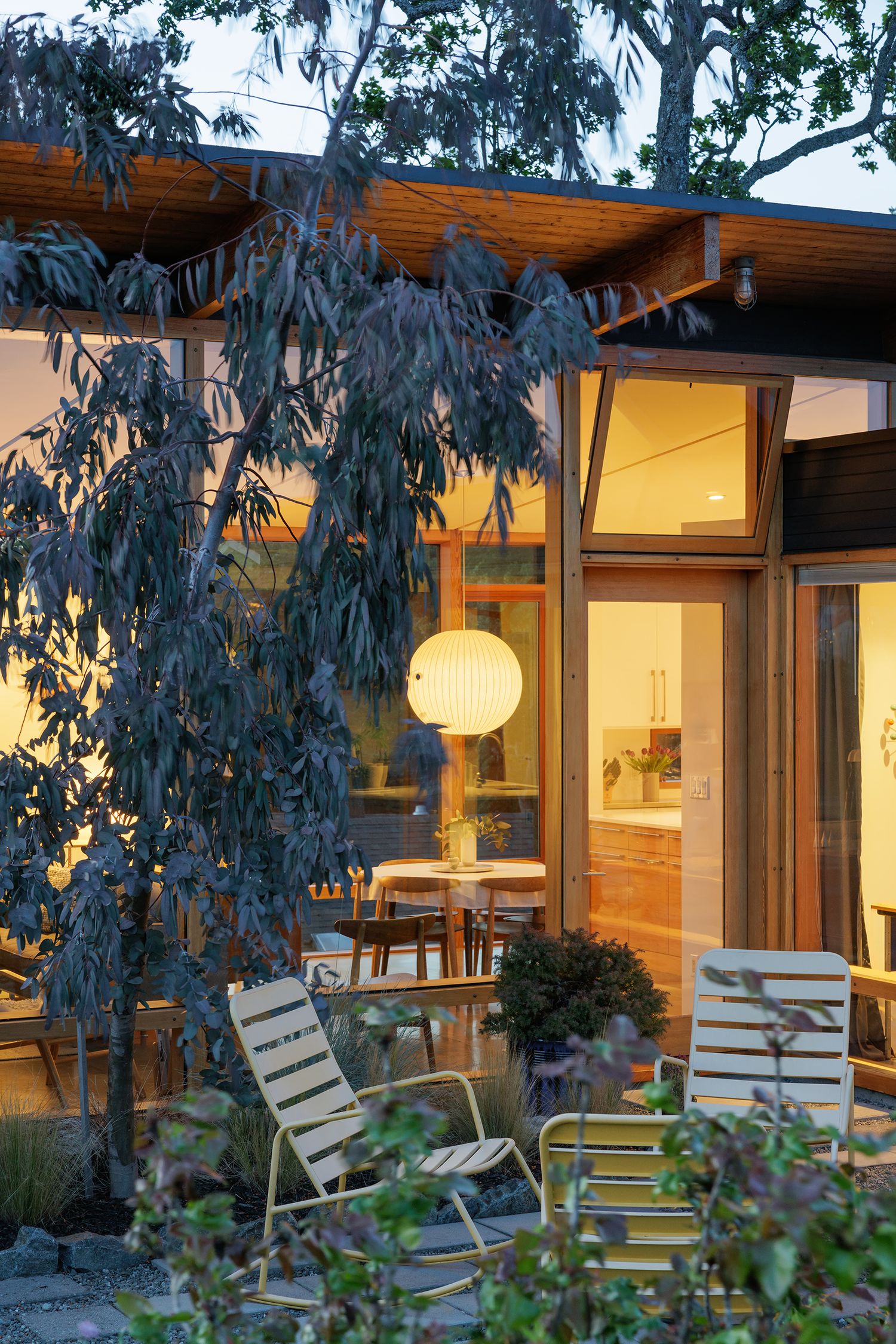 Contemporary home exterior at dusk with warm interior lighting, globe pendant, patio chairs, and lush surrounding trees.