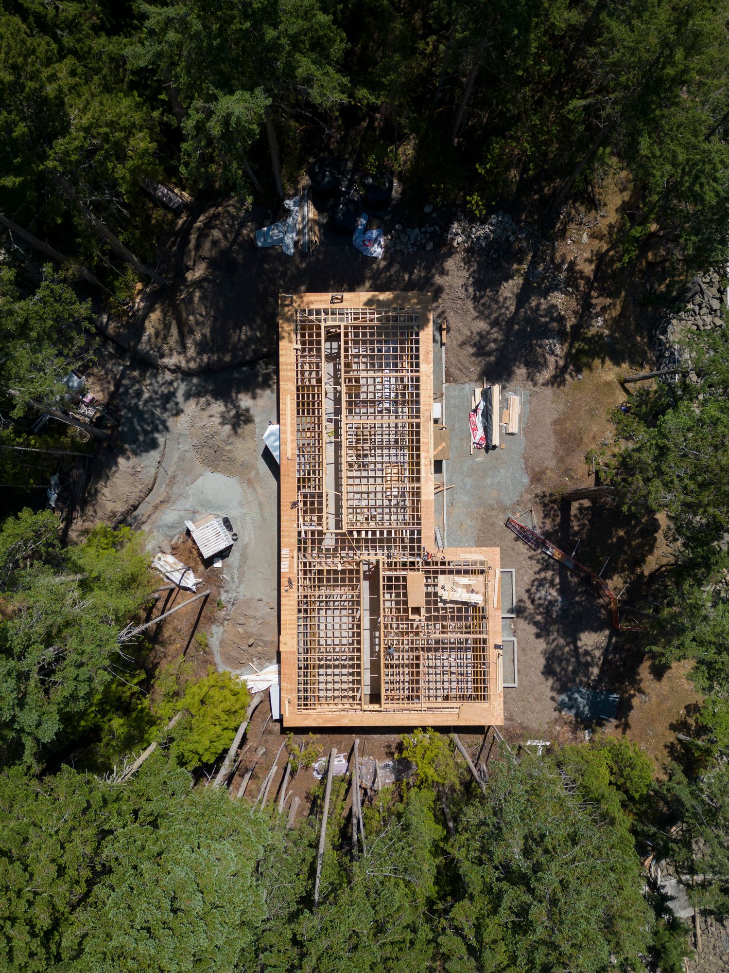 Aerial drone view of timber frame house construction surrounded by tall green forest trees.