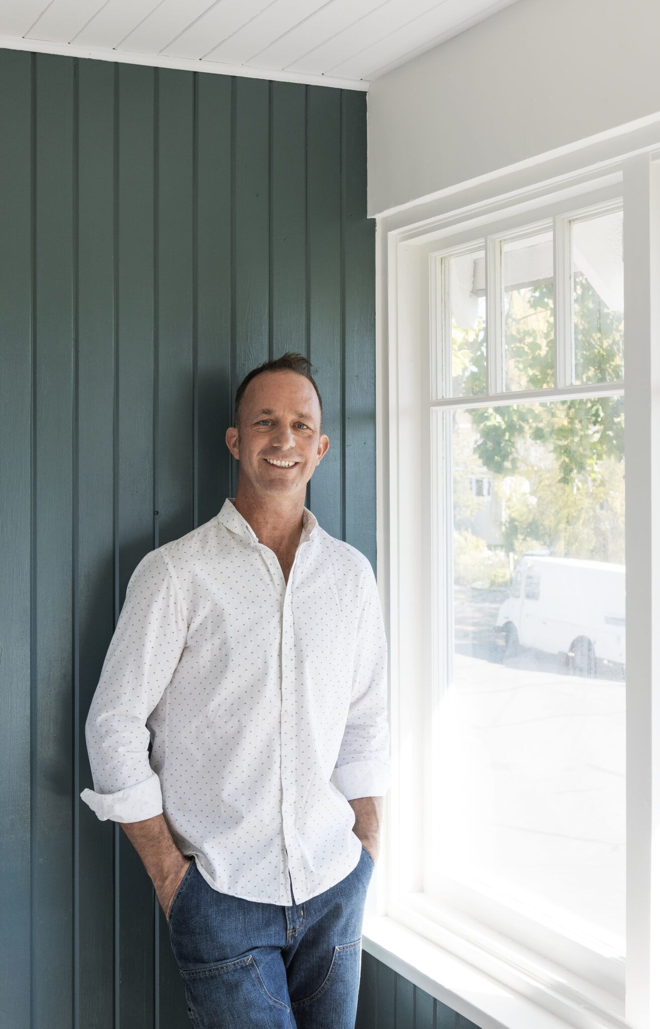 Smiling man in white shirt leaning against dark green board and batten wall beside white window frame.