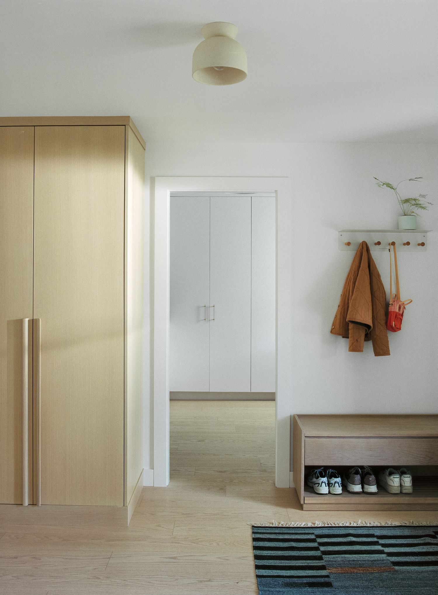 Minimalist entryway with light wood wardrobe, shoe bench, wall hooks with hanging coats, and striped area rug.