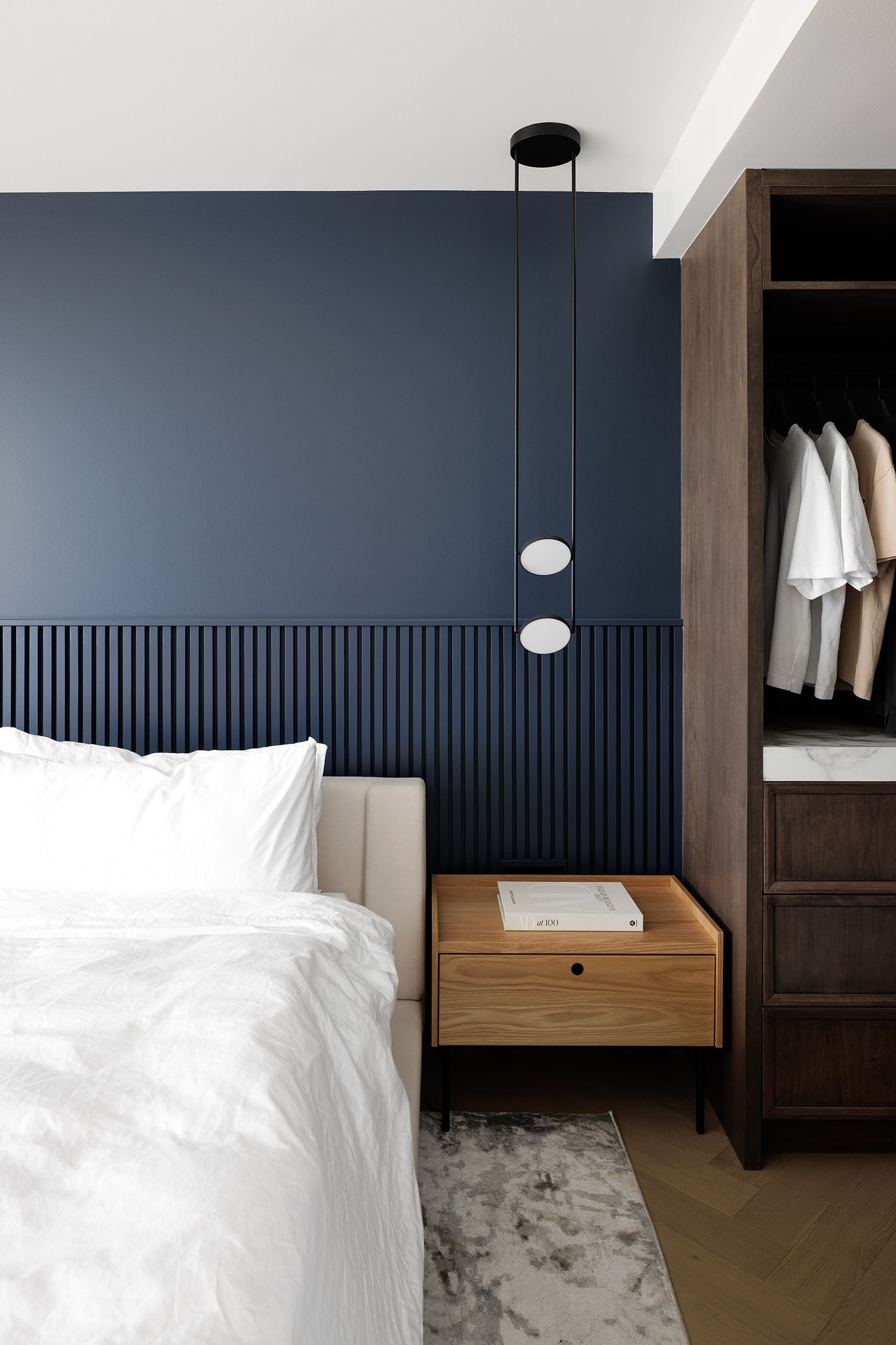 Modern bedroom with navy blue slat accent wall, oak nightstand, pendant light, and open walnut wardrobe.