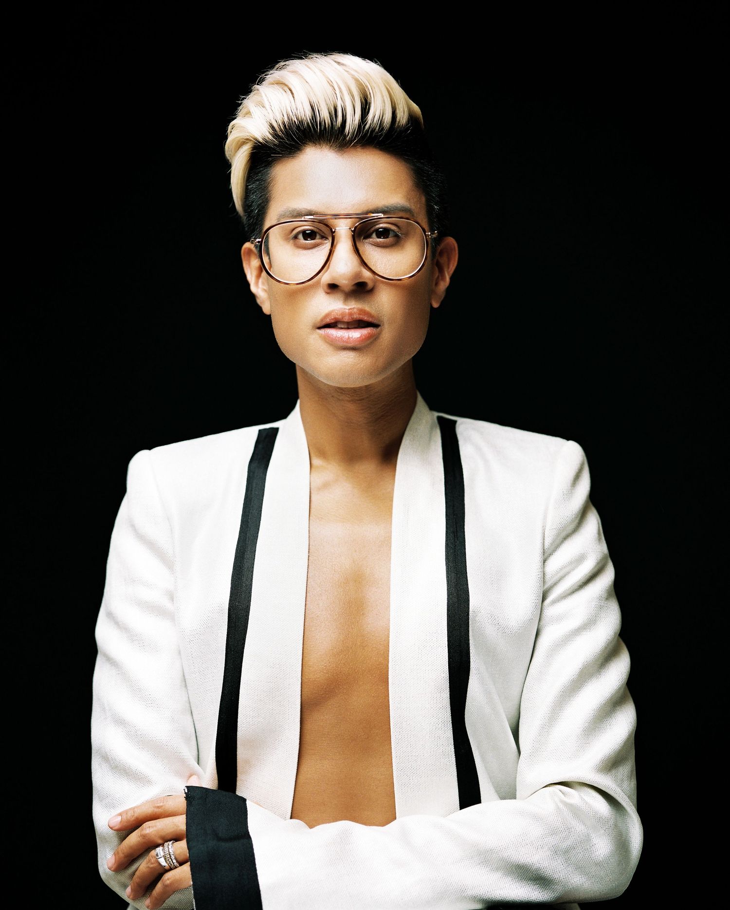 Person with blonde undercut in white blazer and black suspenders posing with arms crossed on black background.