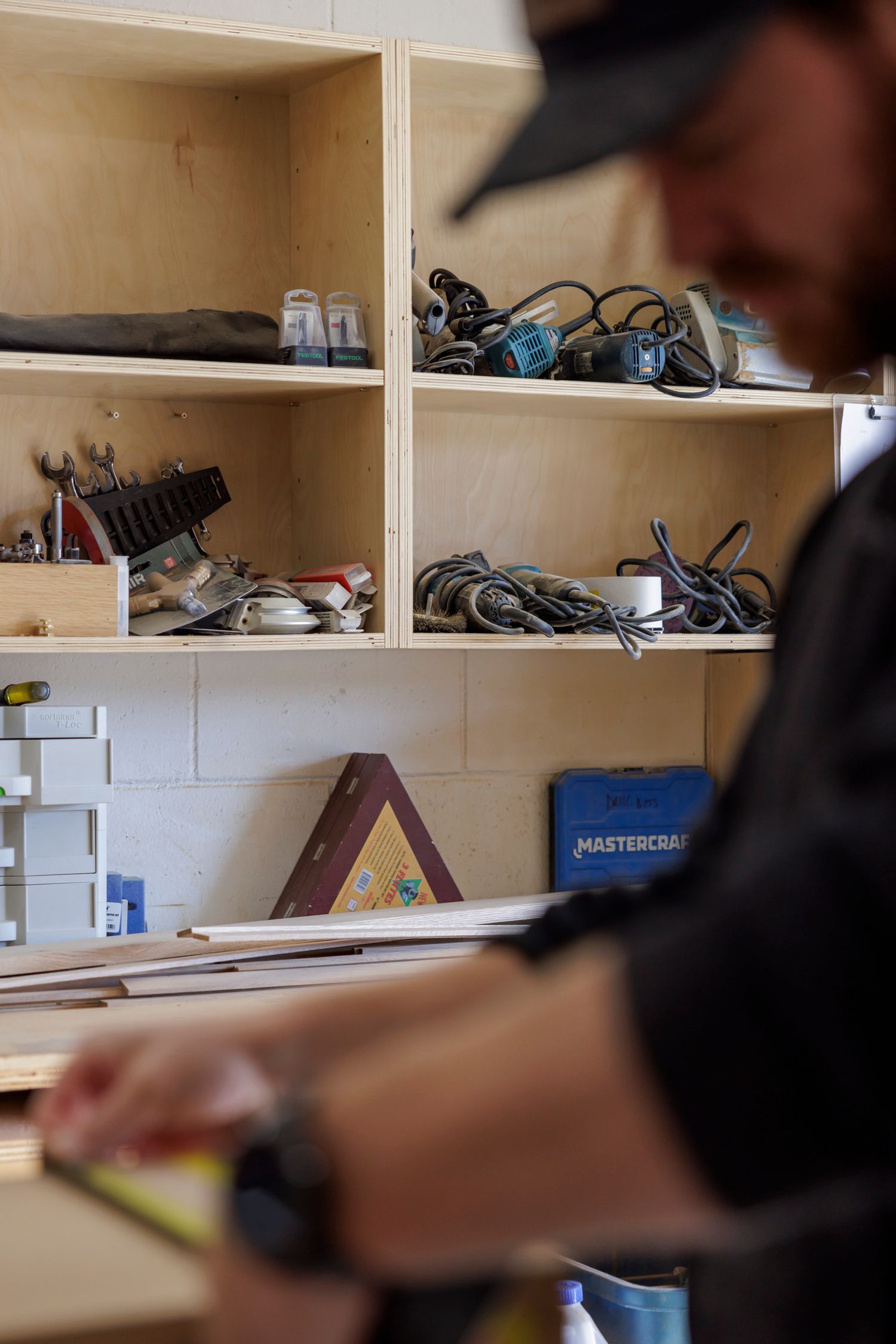 Blurred craftsman measuring wood in workshop with tools and supplies on plywood shelving.