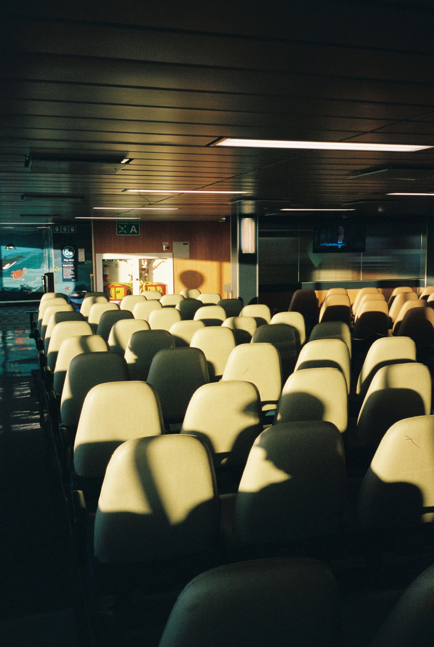 Empty white passenger seats in rows inside a dimly lit ferry cabin with exit signs visible.