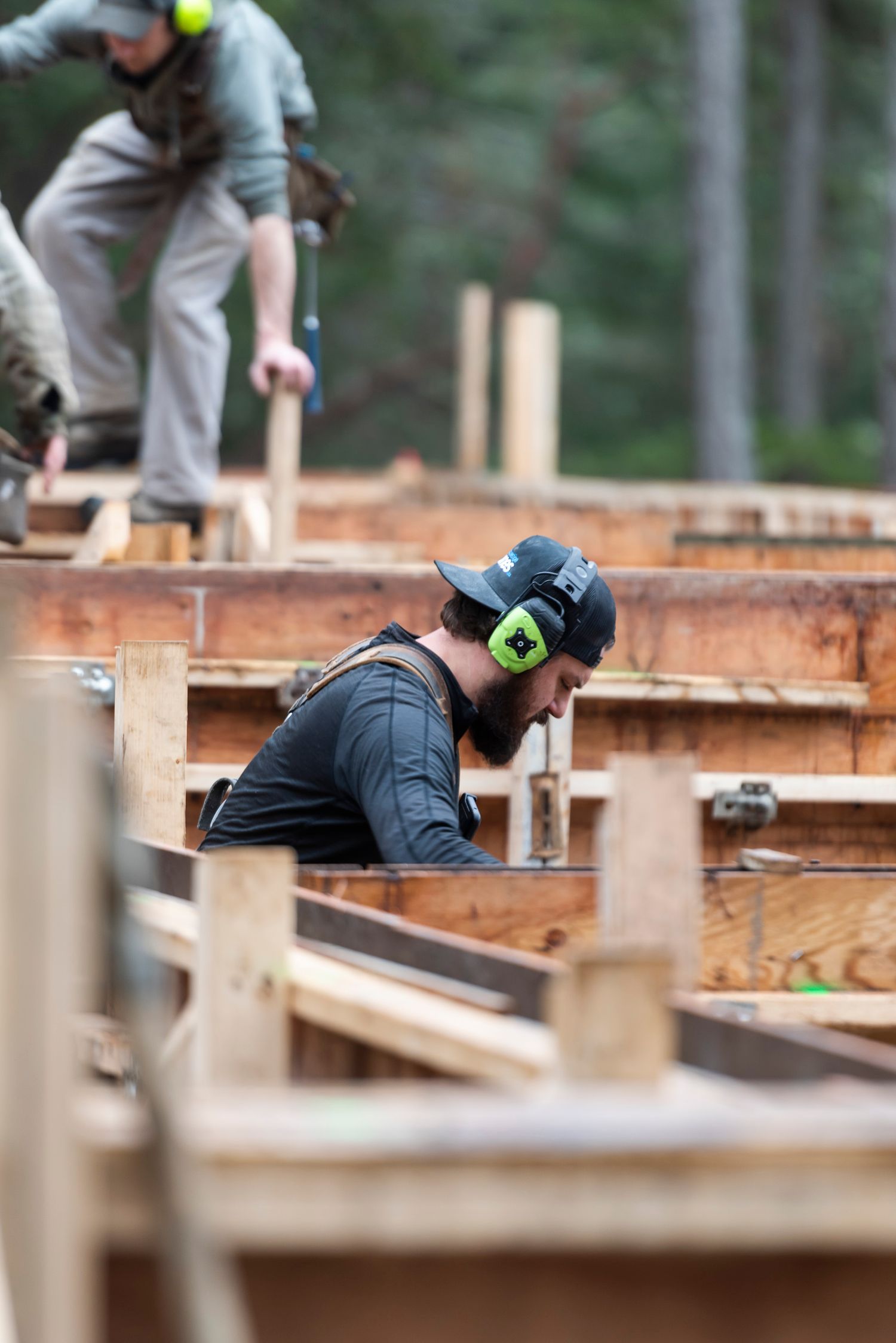 Bearded construction worker wearing ear protection and hard hat working on wooden framework at a building site.