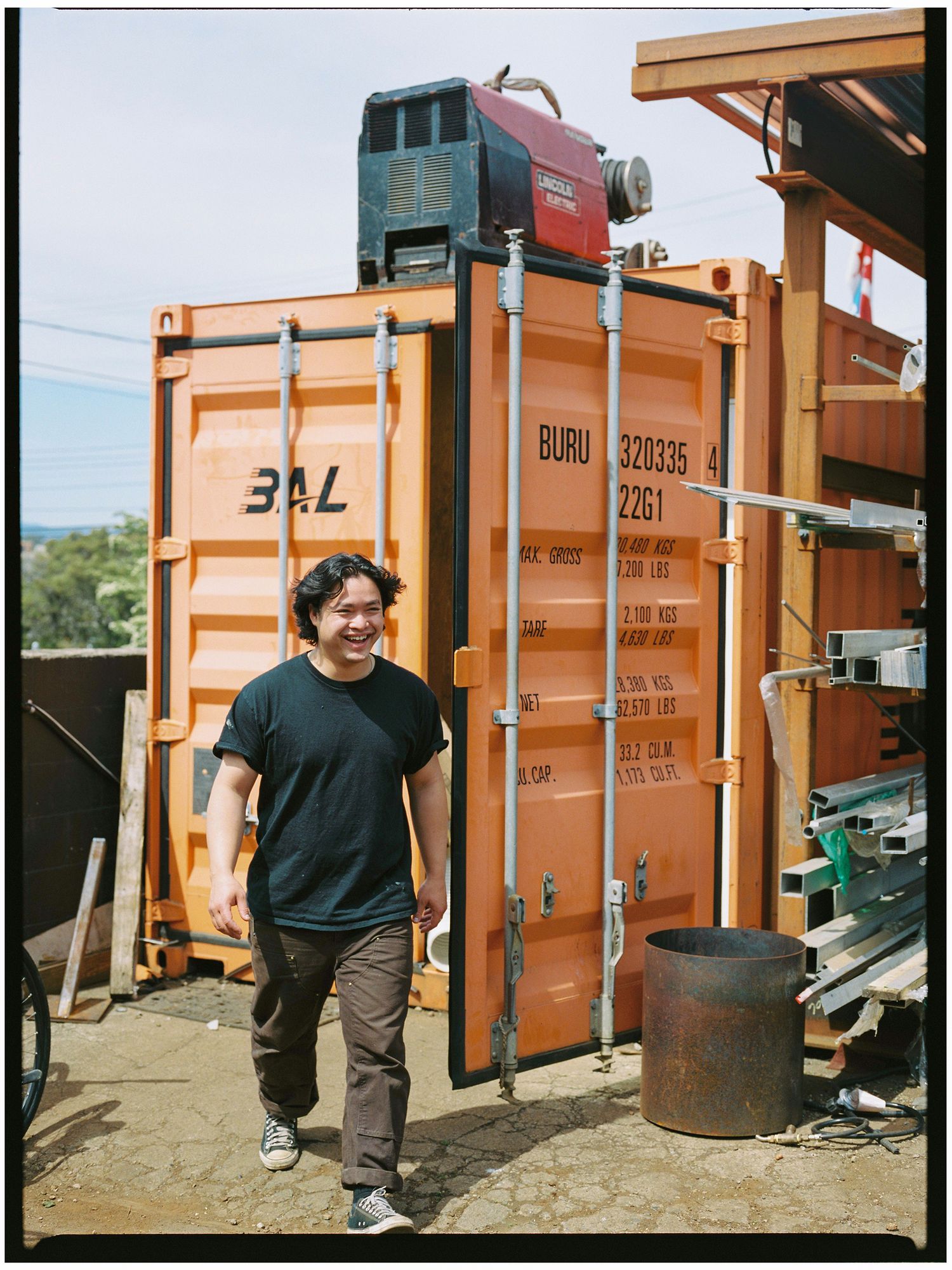 Smiling man walks past an orange BAL shipping container at an outdoor worksite.