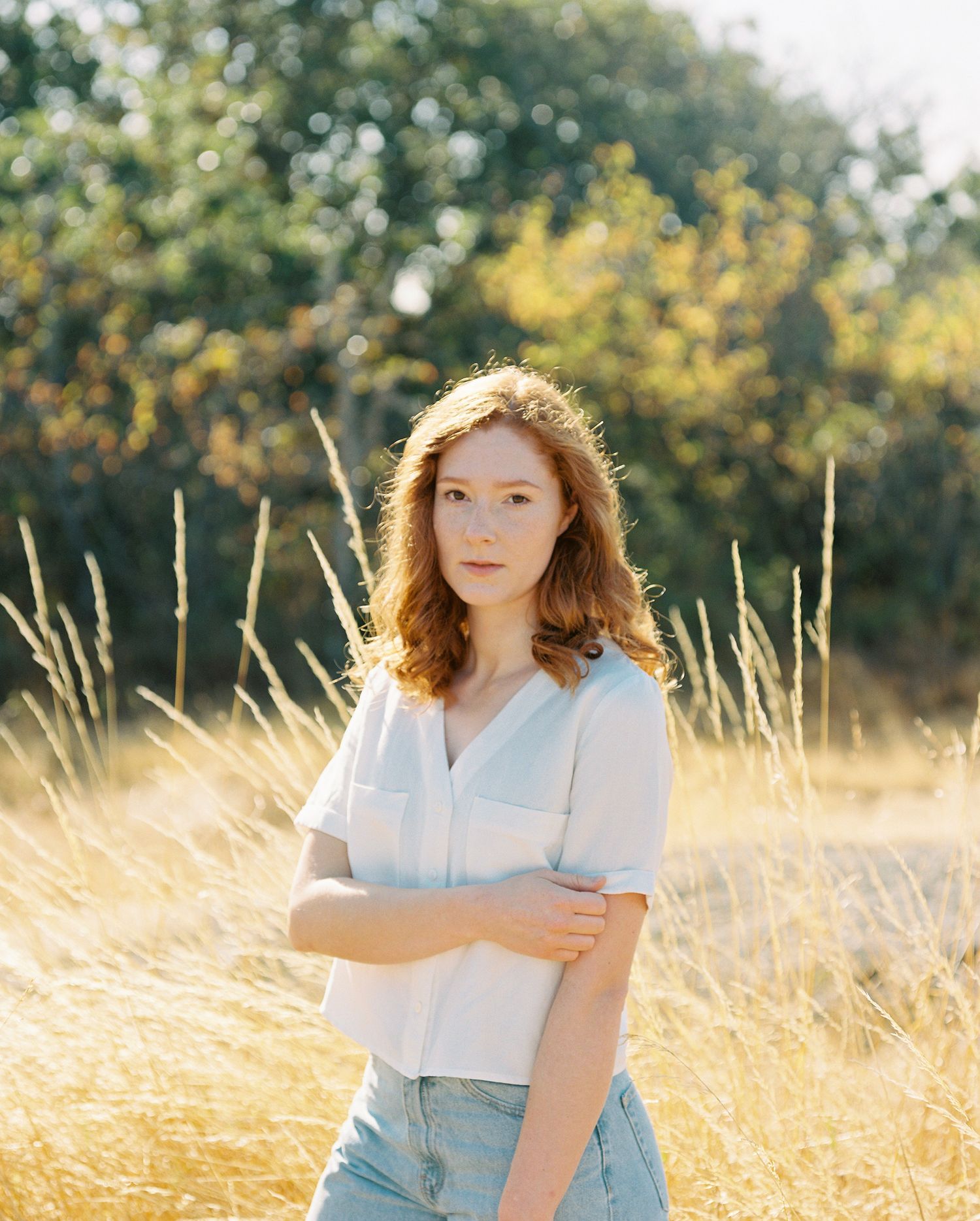 Young redhead woman in white blouse stands in golden sunlit field with trees behind.