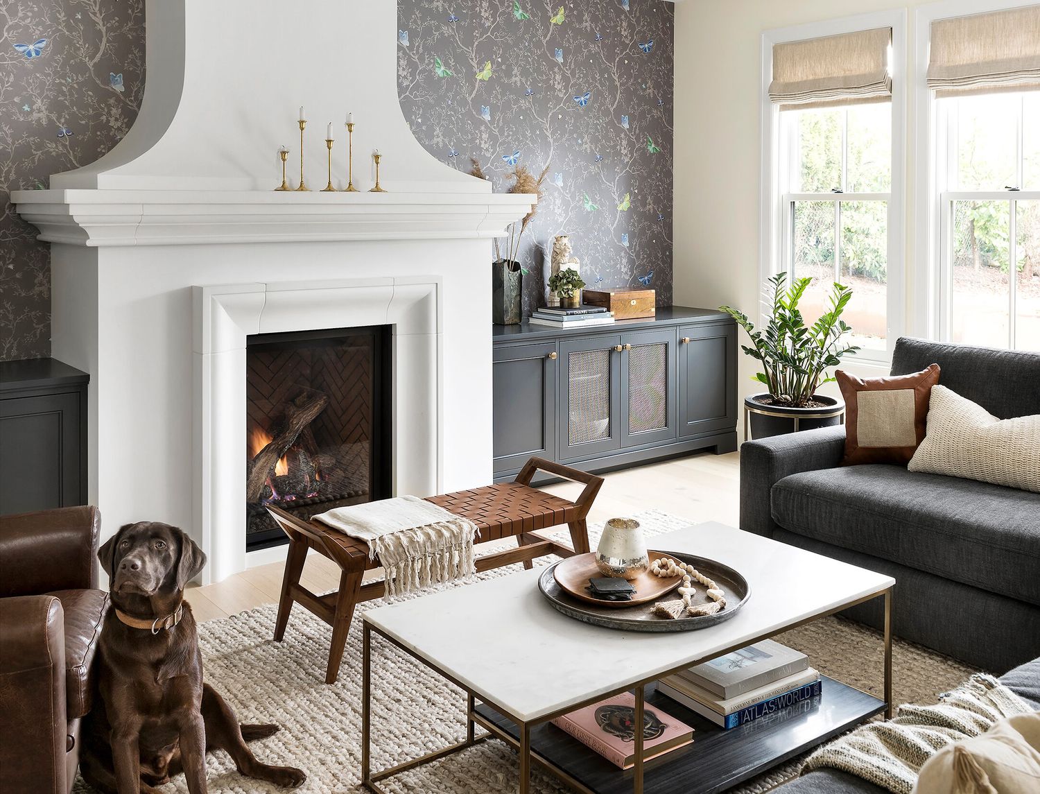Cozy living room with white fireplace, textured wallpaper, dark sofa, leather chair, and a chocolate lab dog sitting nearby.