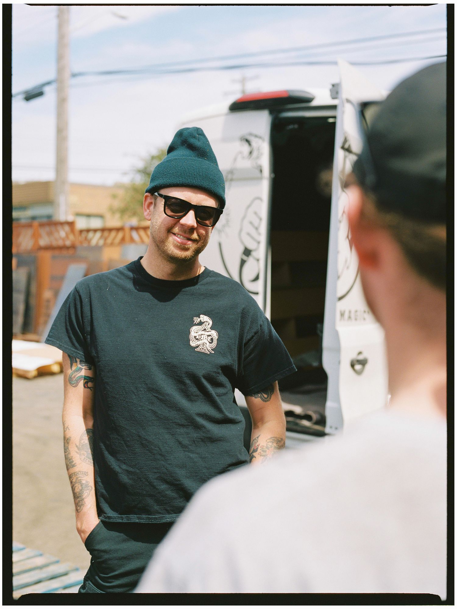 Tattooed man in dark beanie and graphic tee smiles outdoors near a work truck.