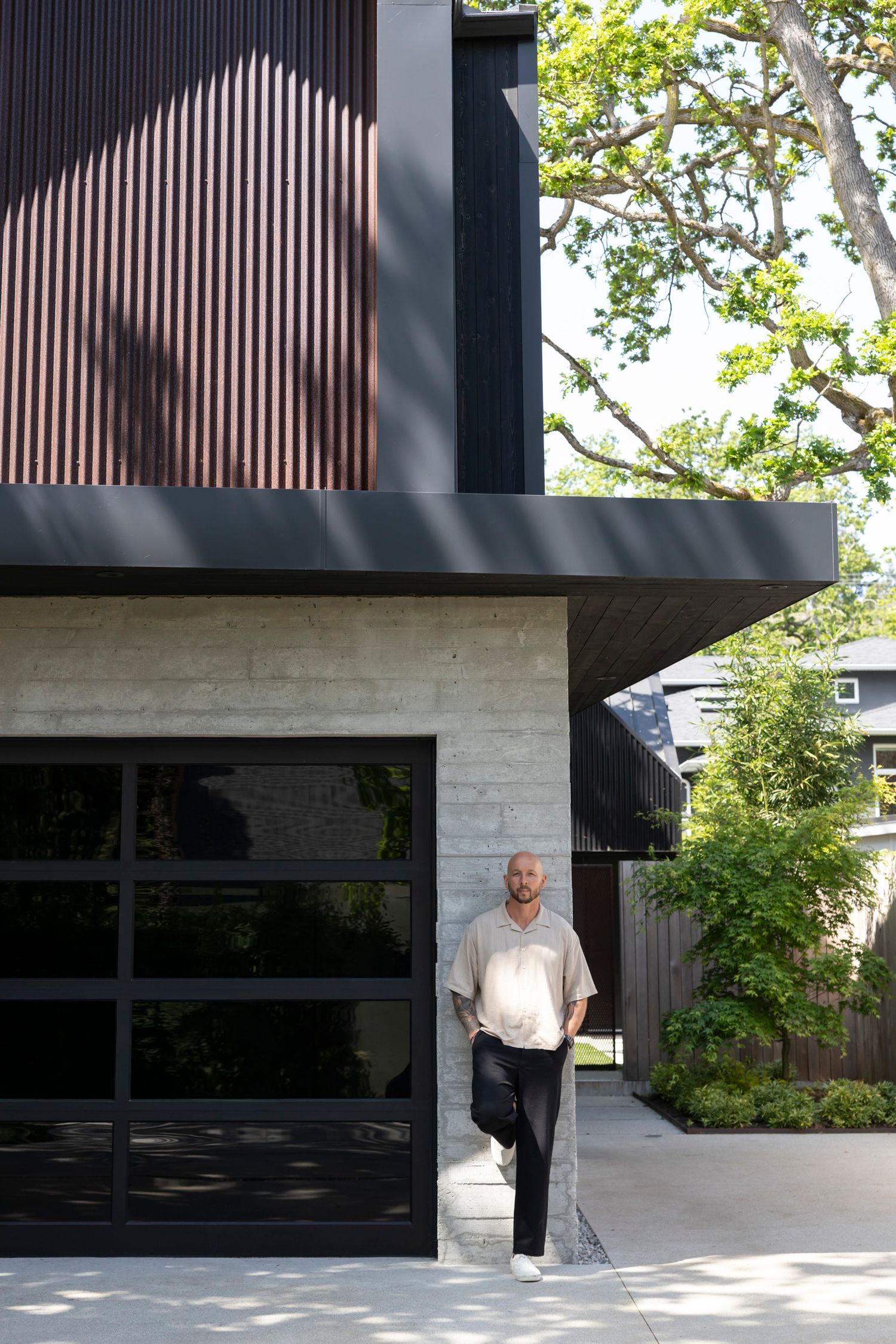 Man in casual shirt stands beside glass garage door of contemporary dark modern home.