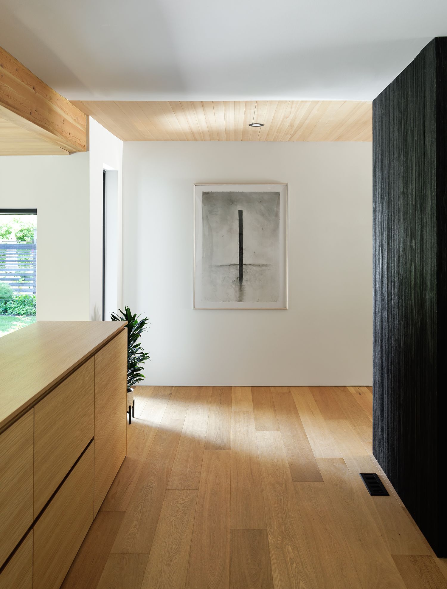 Minimalist interior with light oak floors, wood ceiling, natural cabinetry, and abstract artwork on white wall.