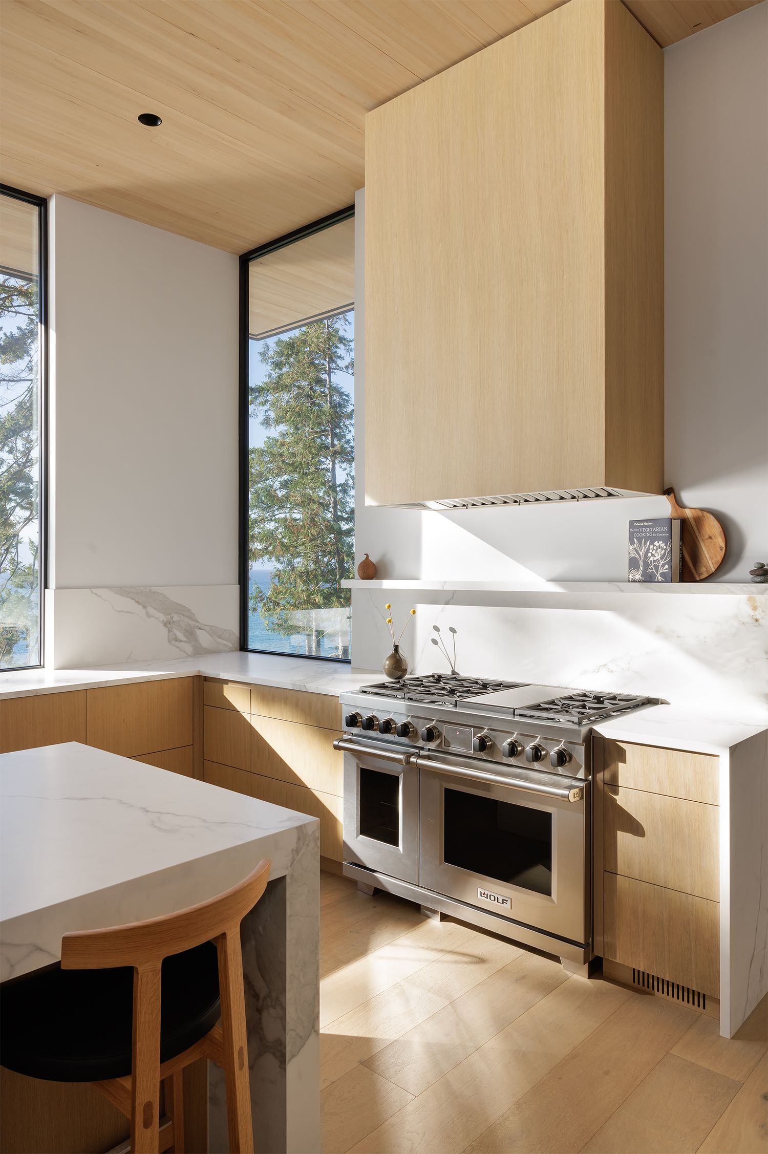 Modern kitchen with light wood cabinetry, white marble countertops, stainless steel Wolf range, and large windows with forest views.