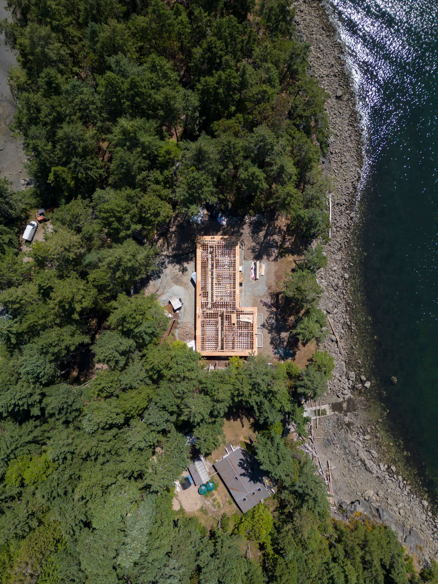 Aerial view of a waterfront home construction site surrounded by lush green forest trees beside a rocky shoreline.