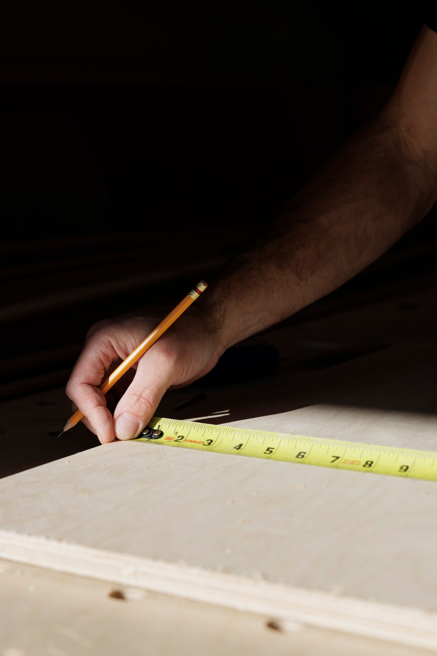 Close-up of hand using pencil and yellow tape measure to mark measurements on light-colored wood panel.