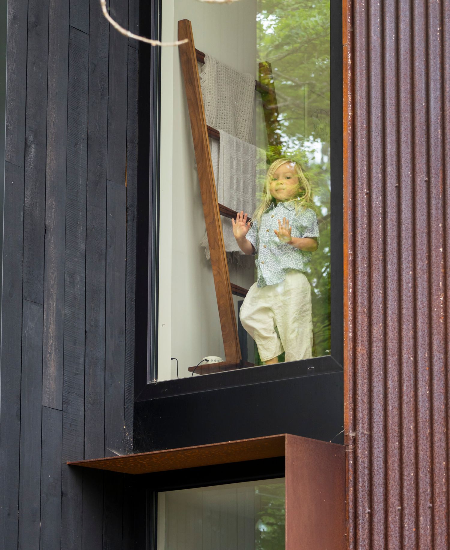 A lifelike child mannequin stands in a window of a modern dark wood building.