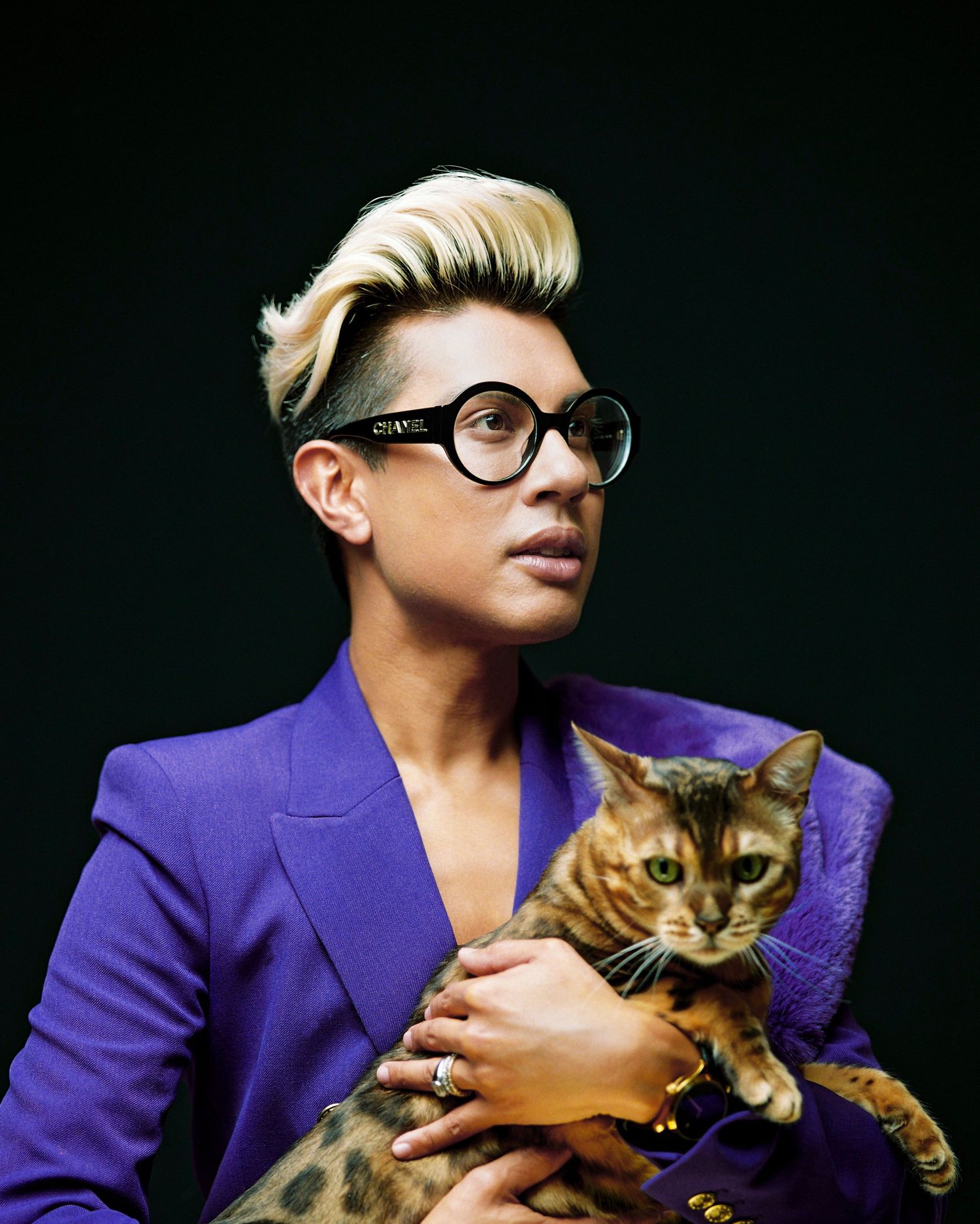 Person with blonde undercut in purple blazer holding a Bengal cat while wearing round Chanel glasses.