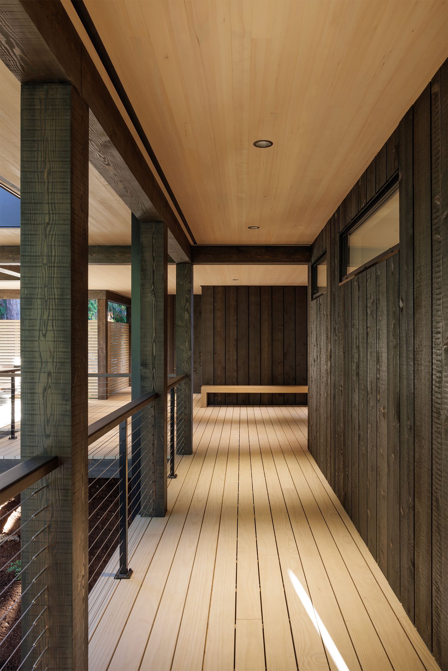 Contemporary covered walkway featuring dark wood siding, light wood deck flooring, wood ceiling, and cable railings with forest views.