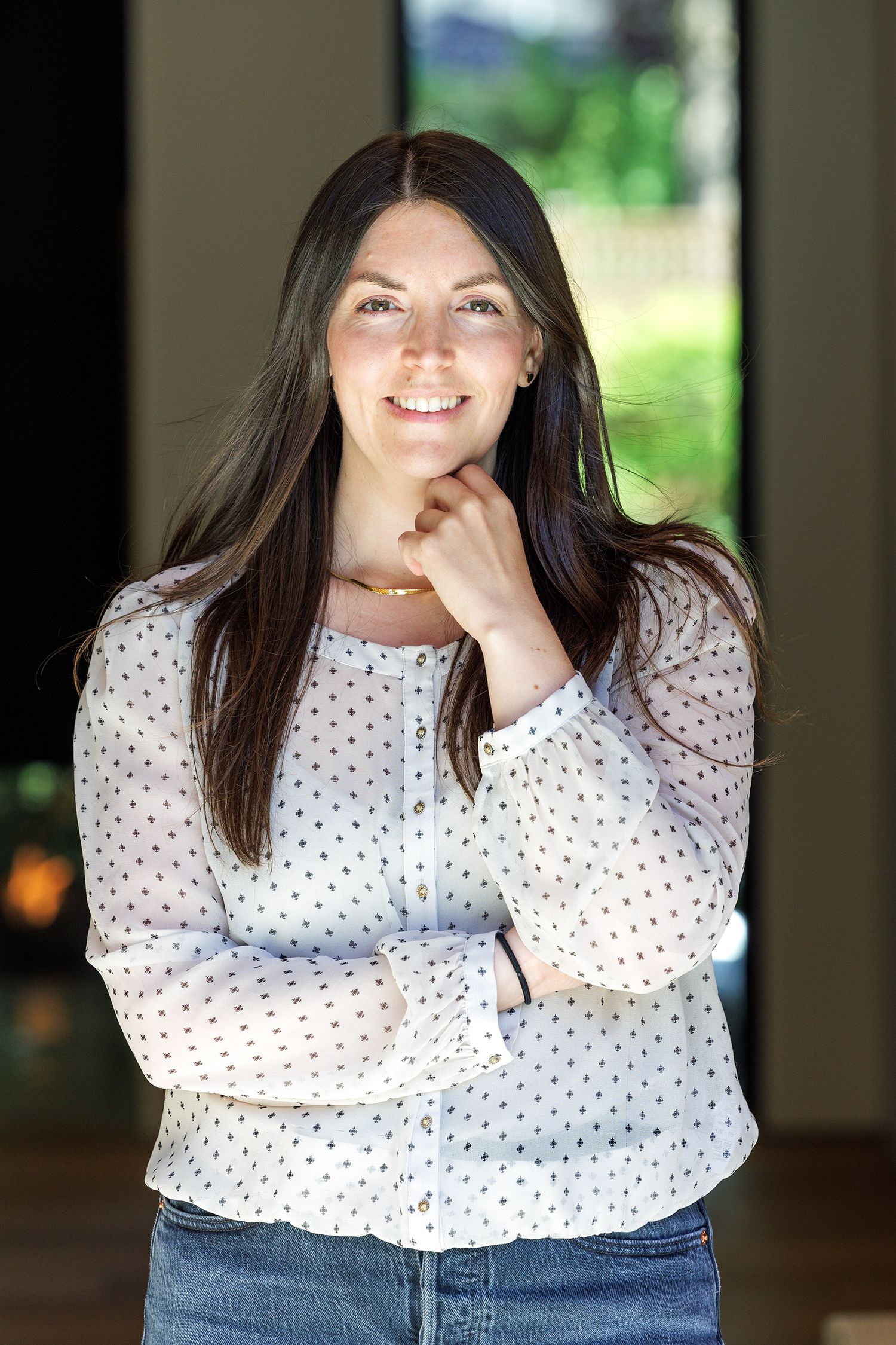 Smiling brunette woman in white polka dot blouse posing thoughtfully with hand on chin in a bright interior.