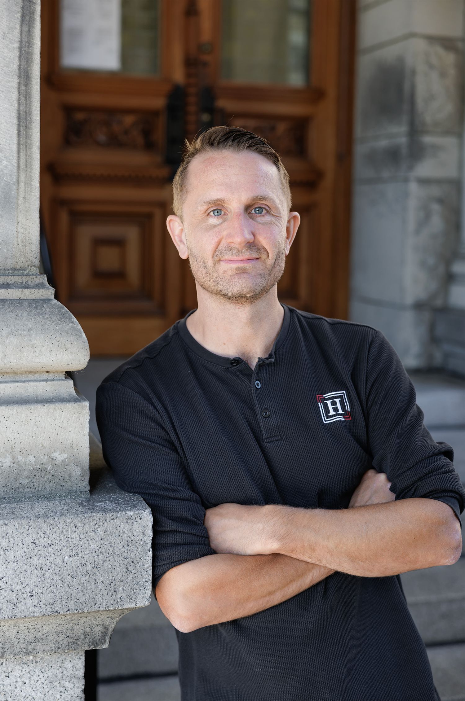 Professional man in black henley shirt standing with arms crossed before ornate wooden doors and stone columns.