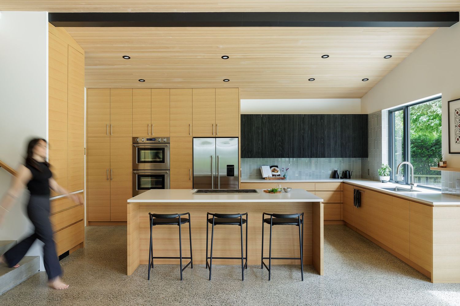 Contemporary open-plan kitchen featuring light wood cabinetry, dark upper cabinets, island with bar stools, and polished concrete floors.