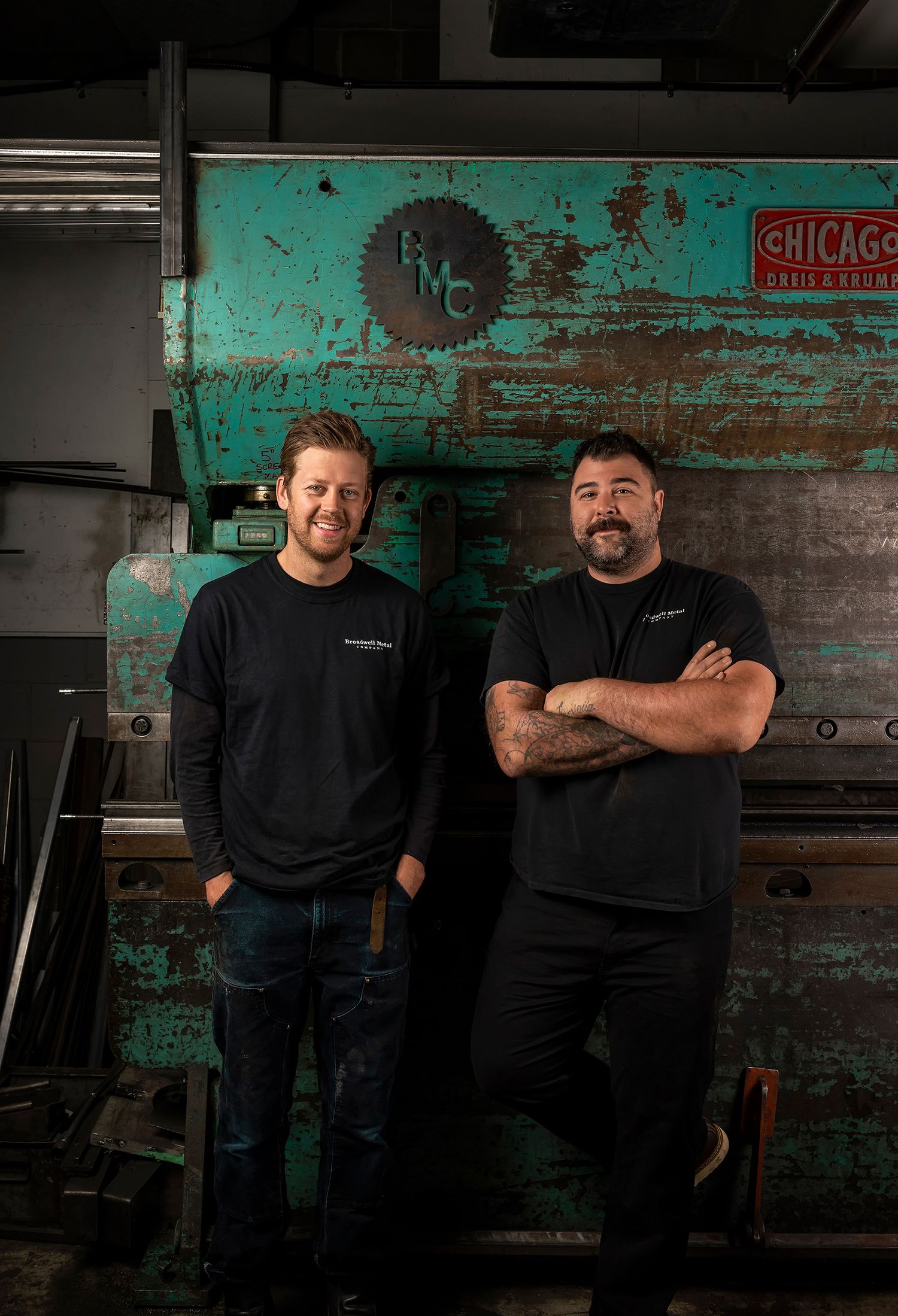 Two men in black shirts posing in front of vintage industrial metalworking equipment in a workshop.