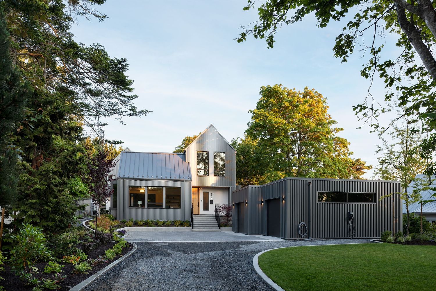 Contemporary home exterior with mixed materials, metal roof, dark cladding, and lush landscaped garden at dusk.