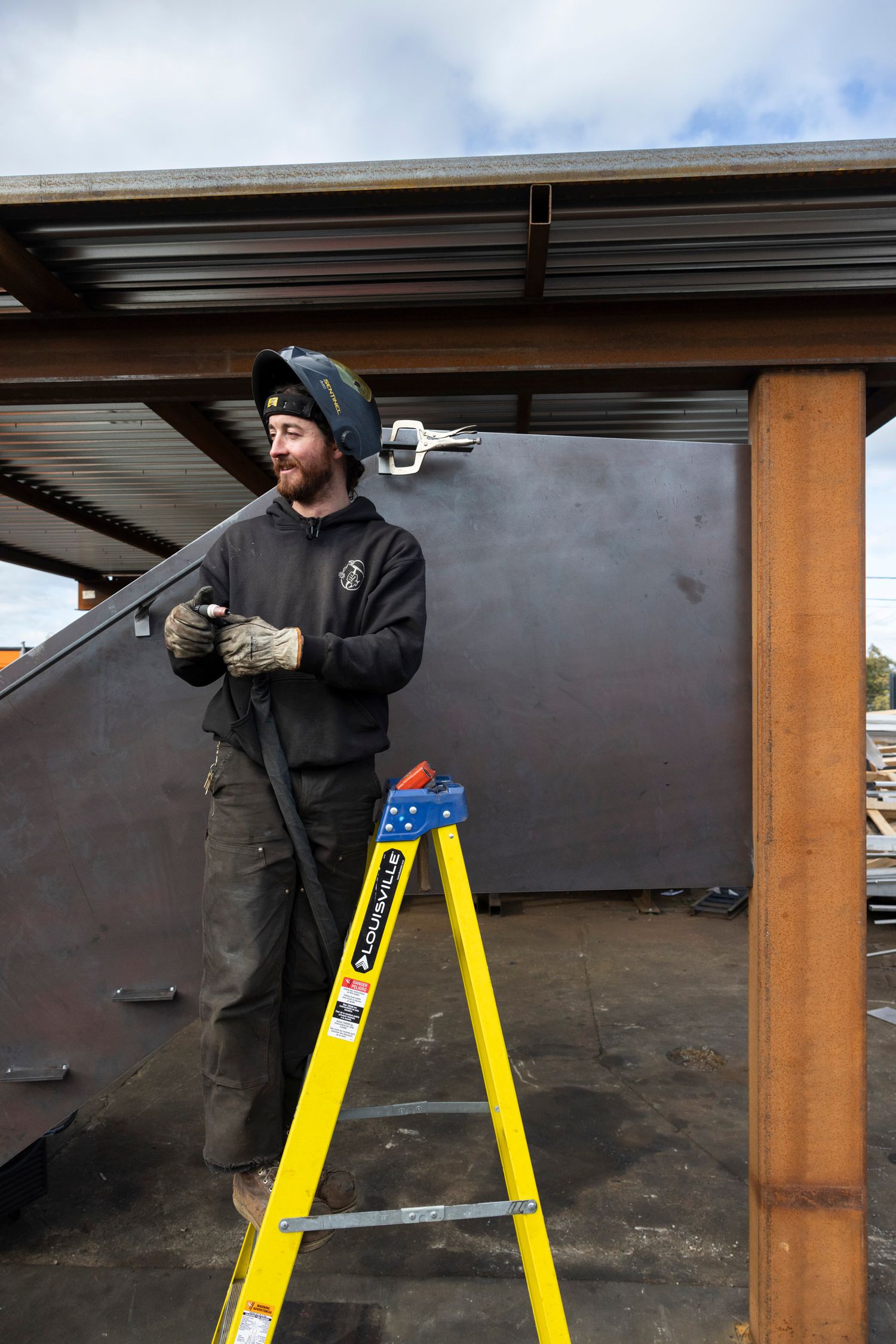 Bearded welder in safety helmet standing on yellow ladder working with large metal panels outdoors.