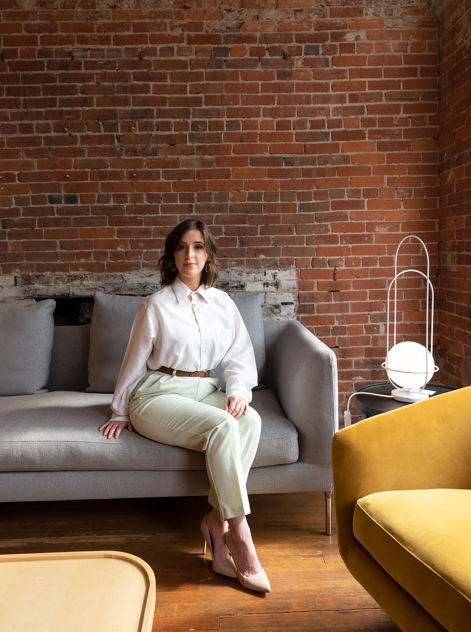 Woman in white blouse and sage pants sitting on grey sofa against exposed brick wall in modern interior.