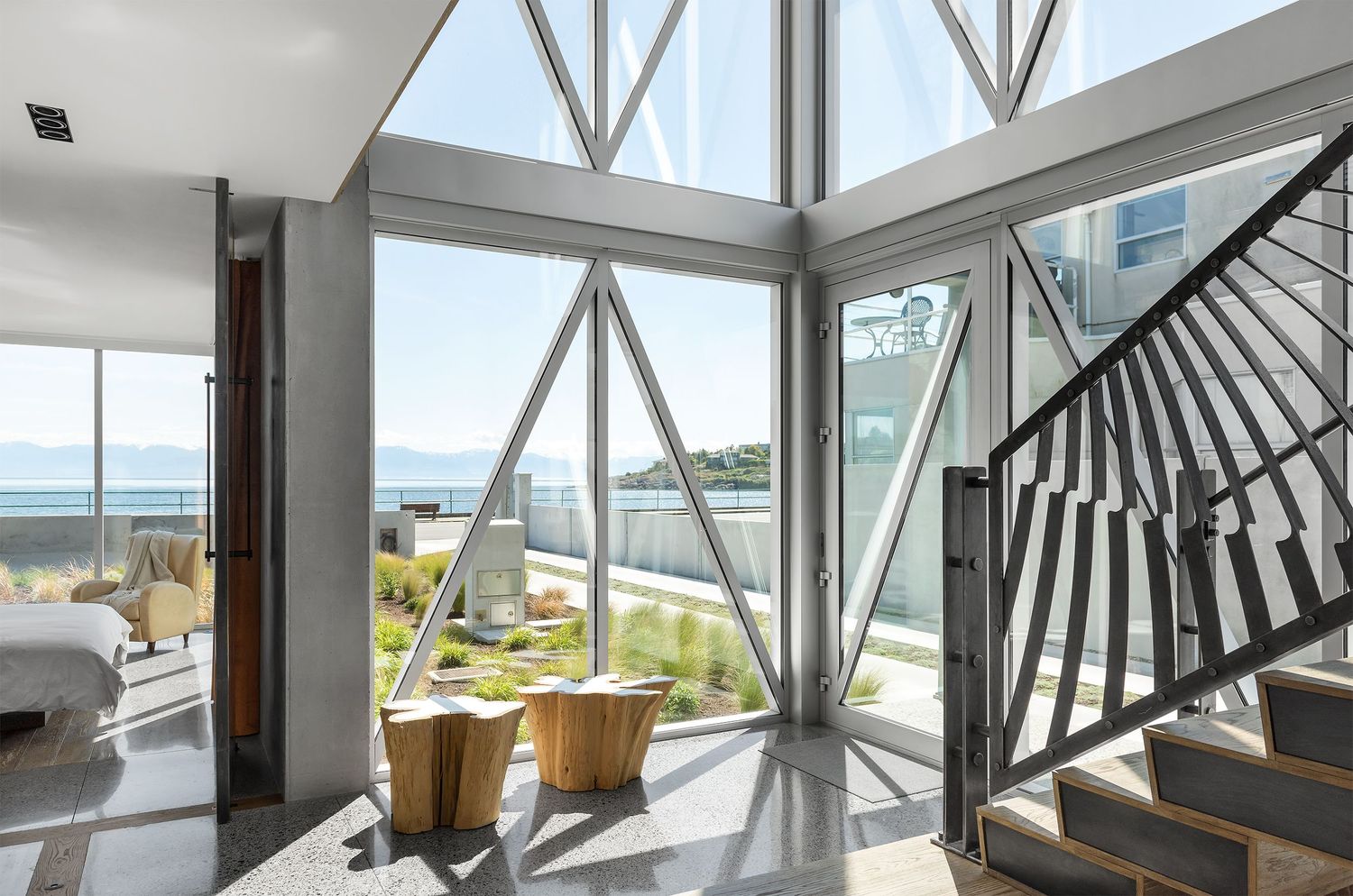 Dramatic entryway with floor-to-ceiling geometric glass windows, natural wood stump stools, and ocean views beyond.