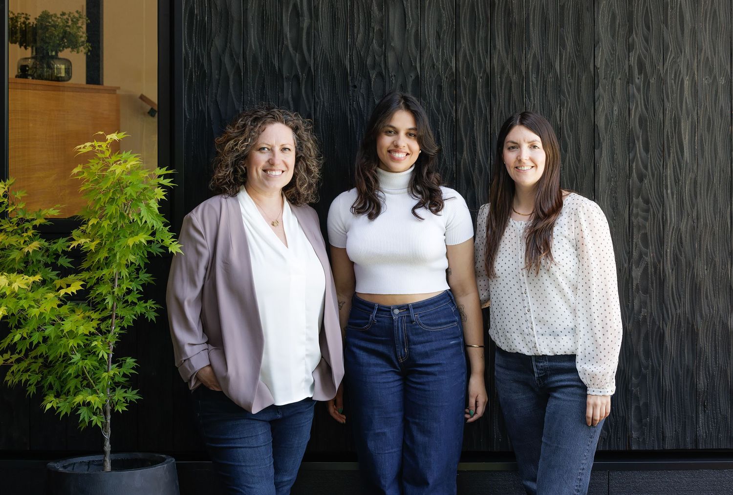 Three smiling women standing together in front of a dark textured wall with green foliage nearby.