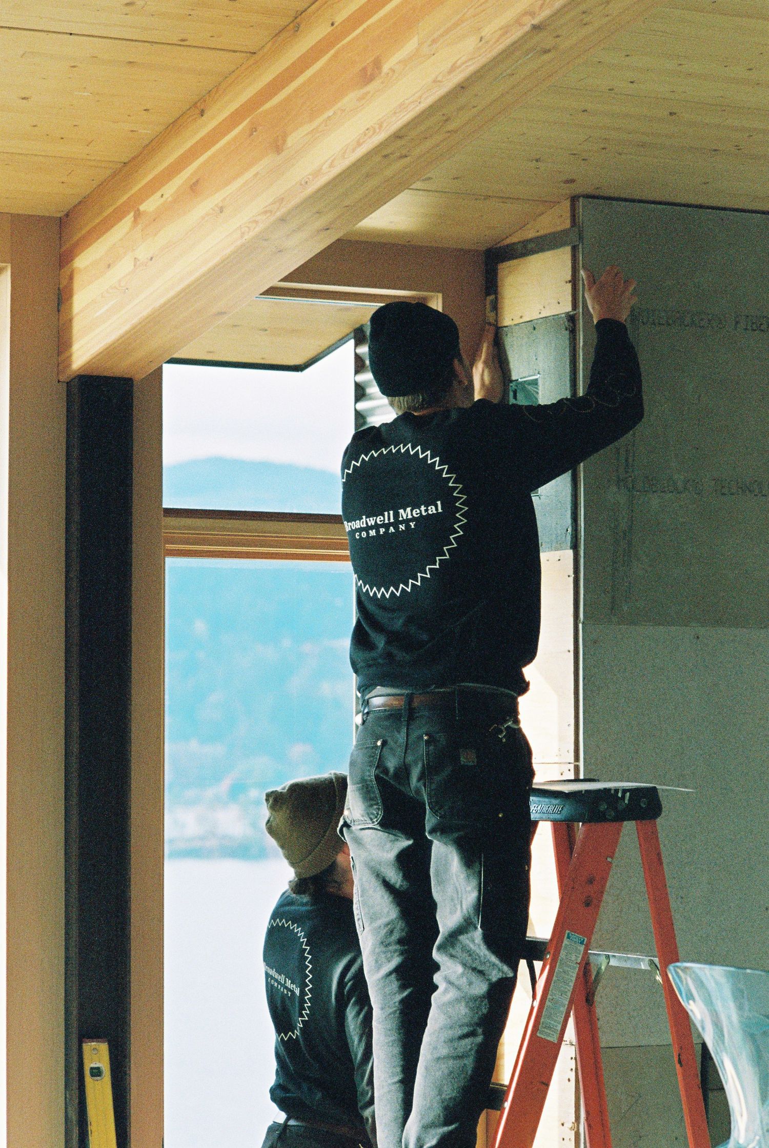 Construction worker standing on a red ladder installs window flashing inside a timber-framed home with ocean views.