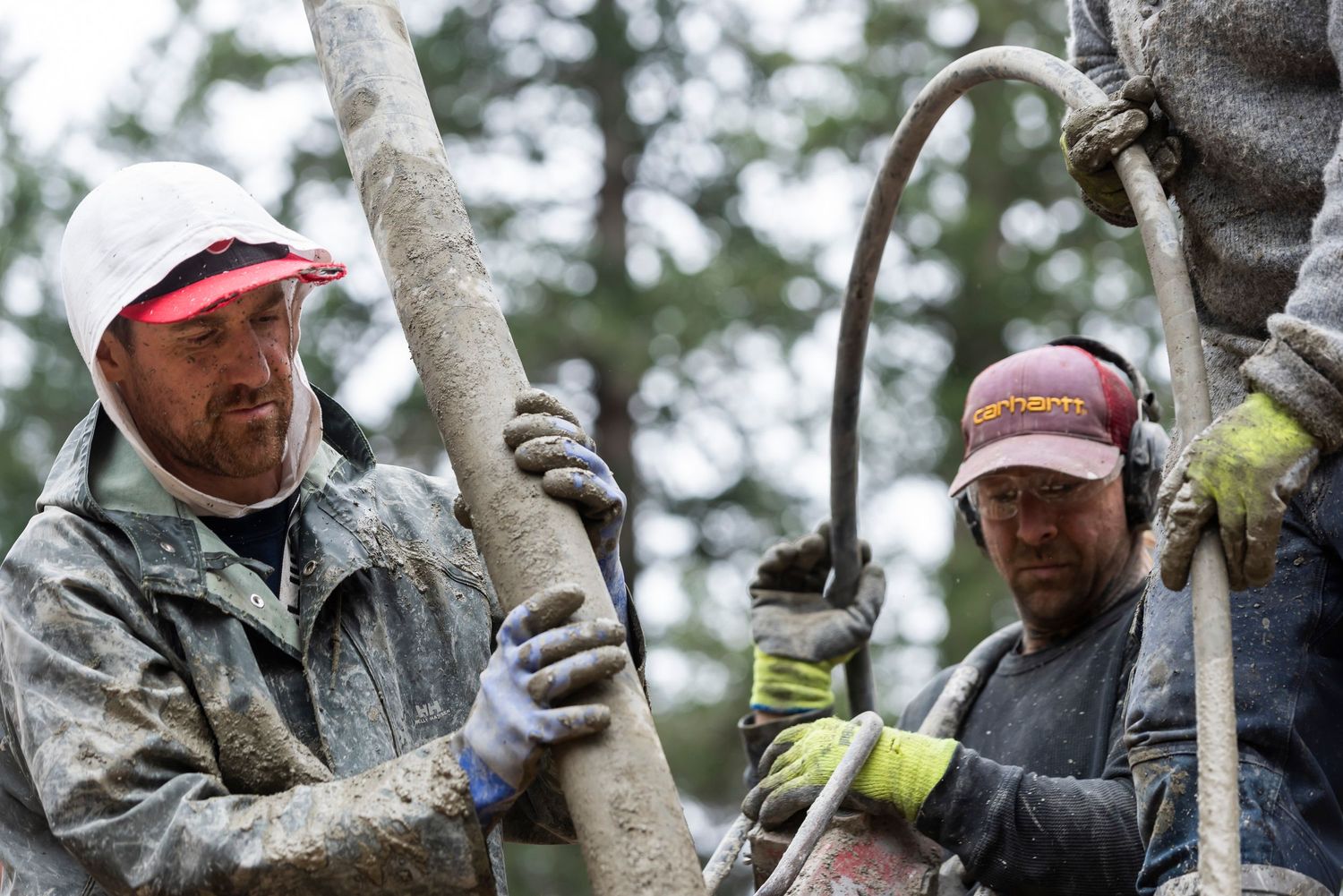 Two workers in caps and gloves handle a concrete pump hose on a muddy outdoor construction site.