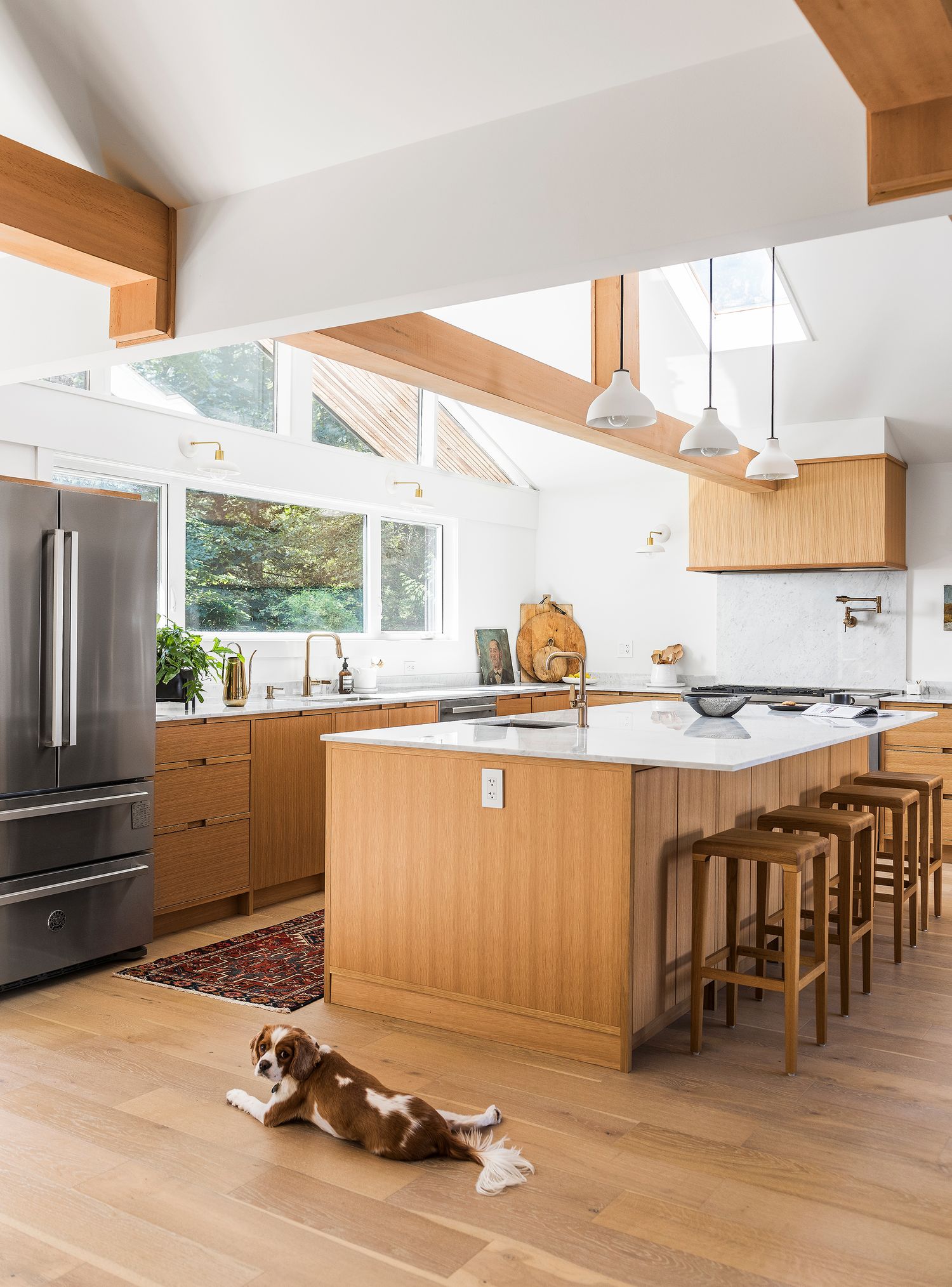 Bright modern kitchen with wood cabinets, marble island, bar stools, vaulted ceiling beams, and a dog lying on the floor.