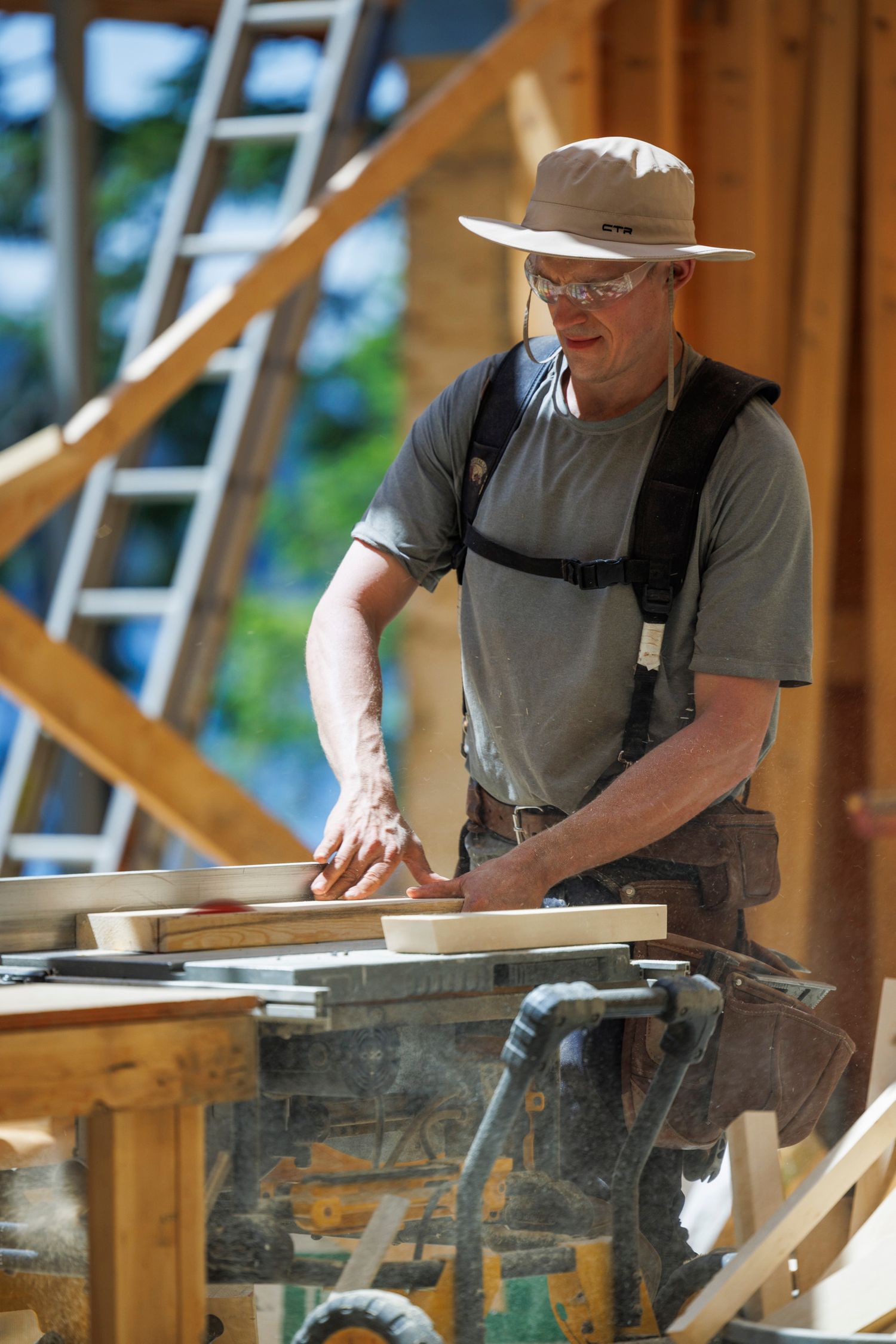 A carpenter in a sun hat and safety glasses cuts wood on a table saw at an outdoor construction site.
