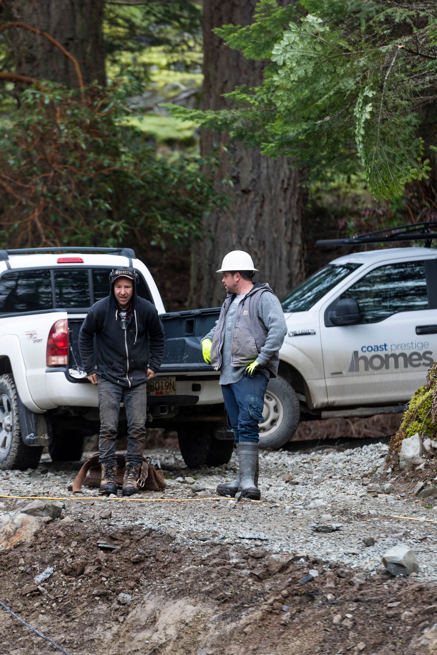 Two construction workers standing near a white Coast Custom Homes pickup truck on a gravel job site.