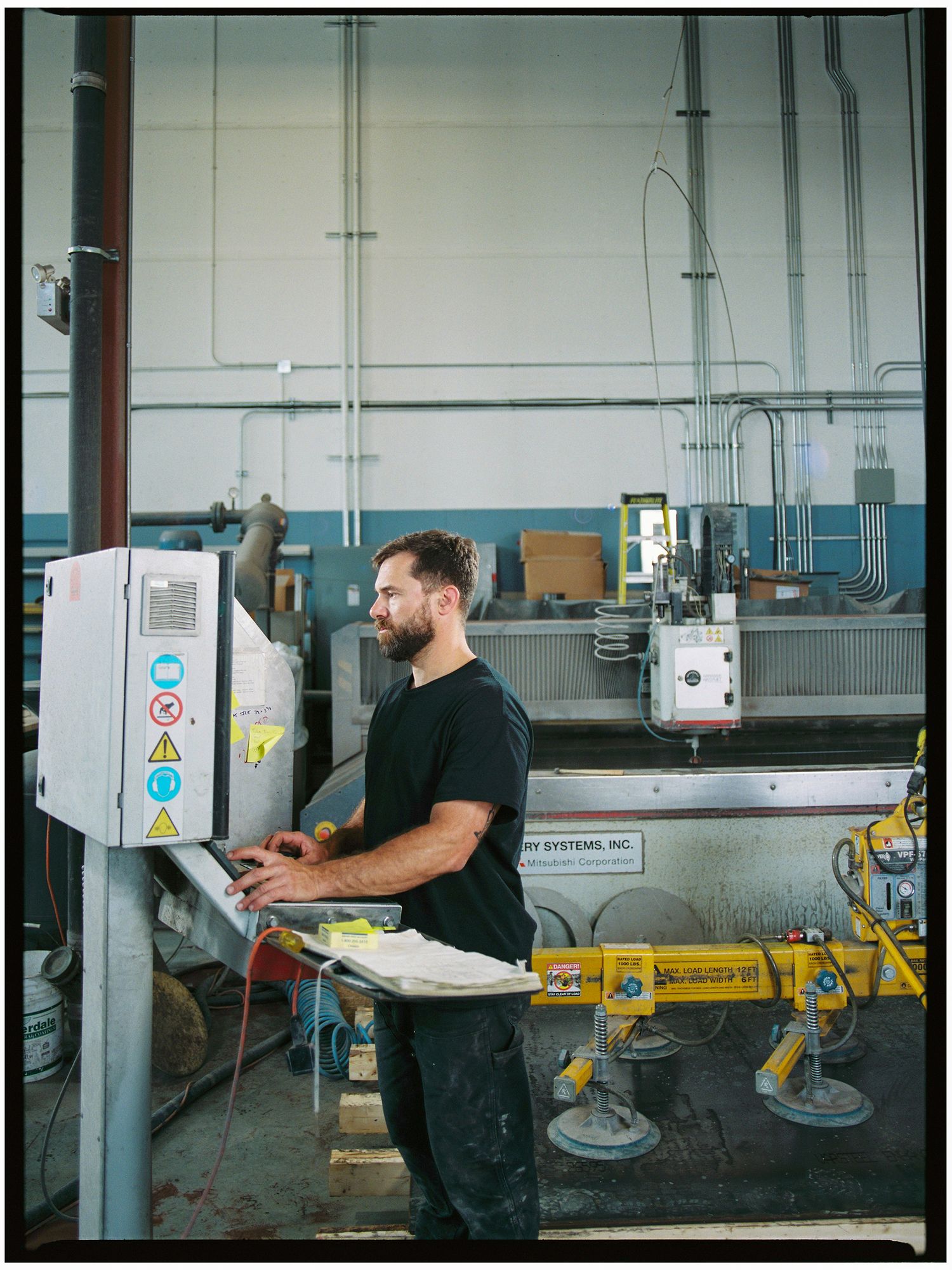 Bearded man operates CNC machinery control panel in an industrial warehouse.