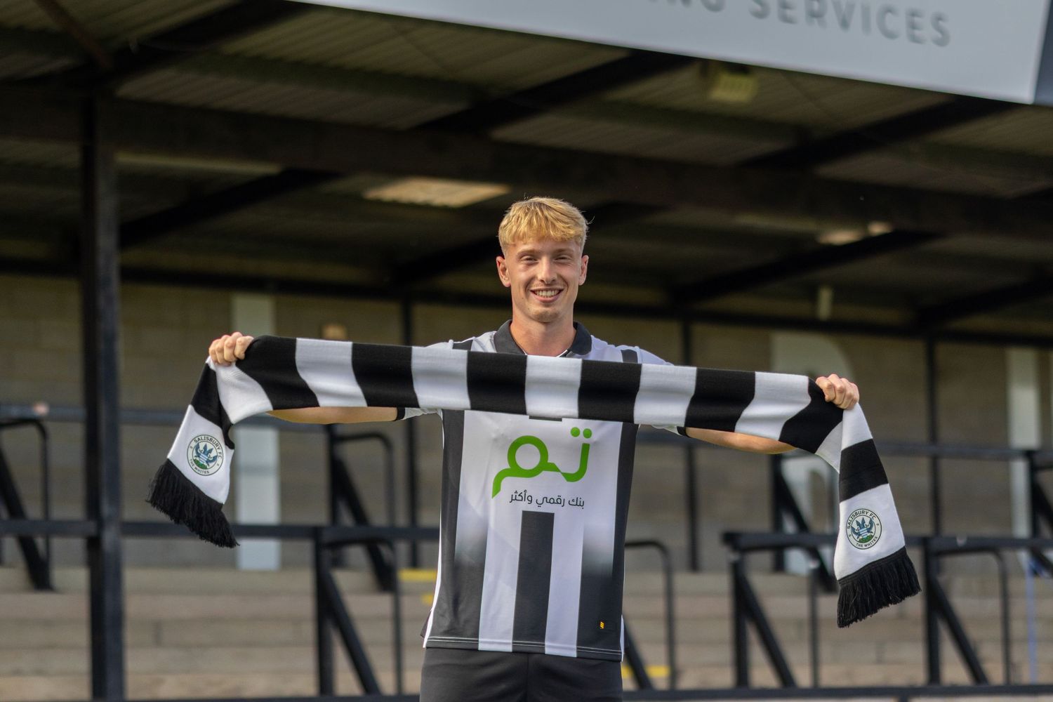 A young blonde soccer player holds up a black and white striped scarf in front of a stadium stand.