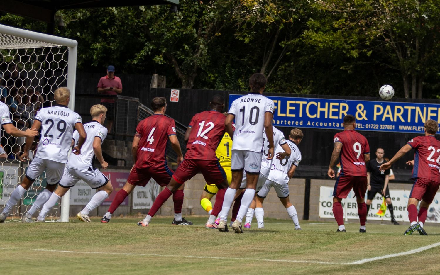 Players in white and maroon kits contest corner kick near goal during sunny football match.