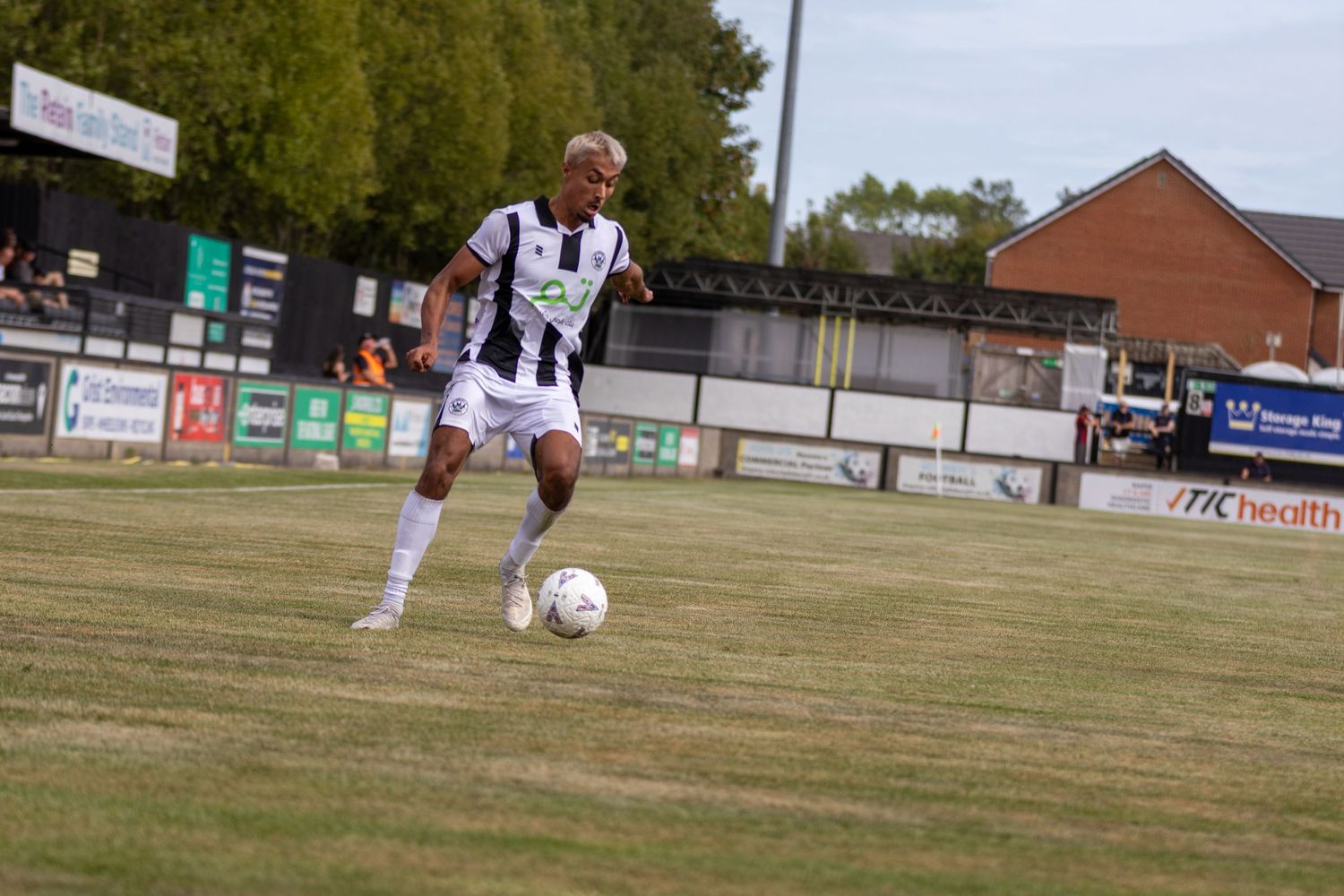 Soccer player in black and white striped kit dribbles ball on grass pitch with stadium stands behind.