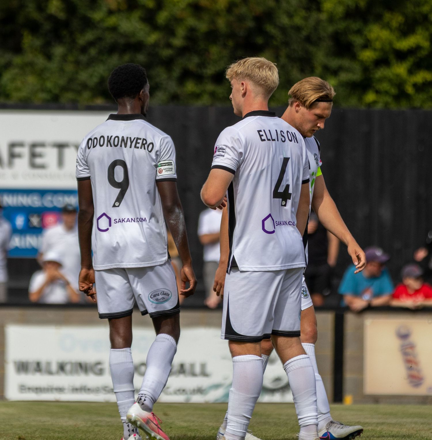 Two footballers named Odokonyero and Ellison in black and white kits walk together on the pitch.