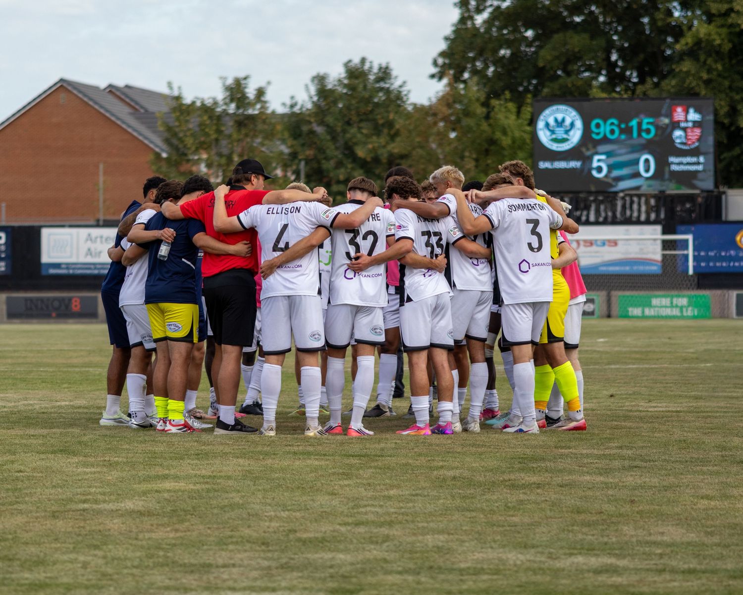 Soccer players in white uniforms huddle together as scoreboard shows 5-0 final score.