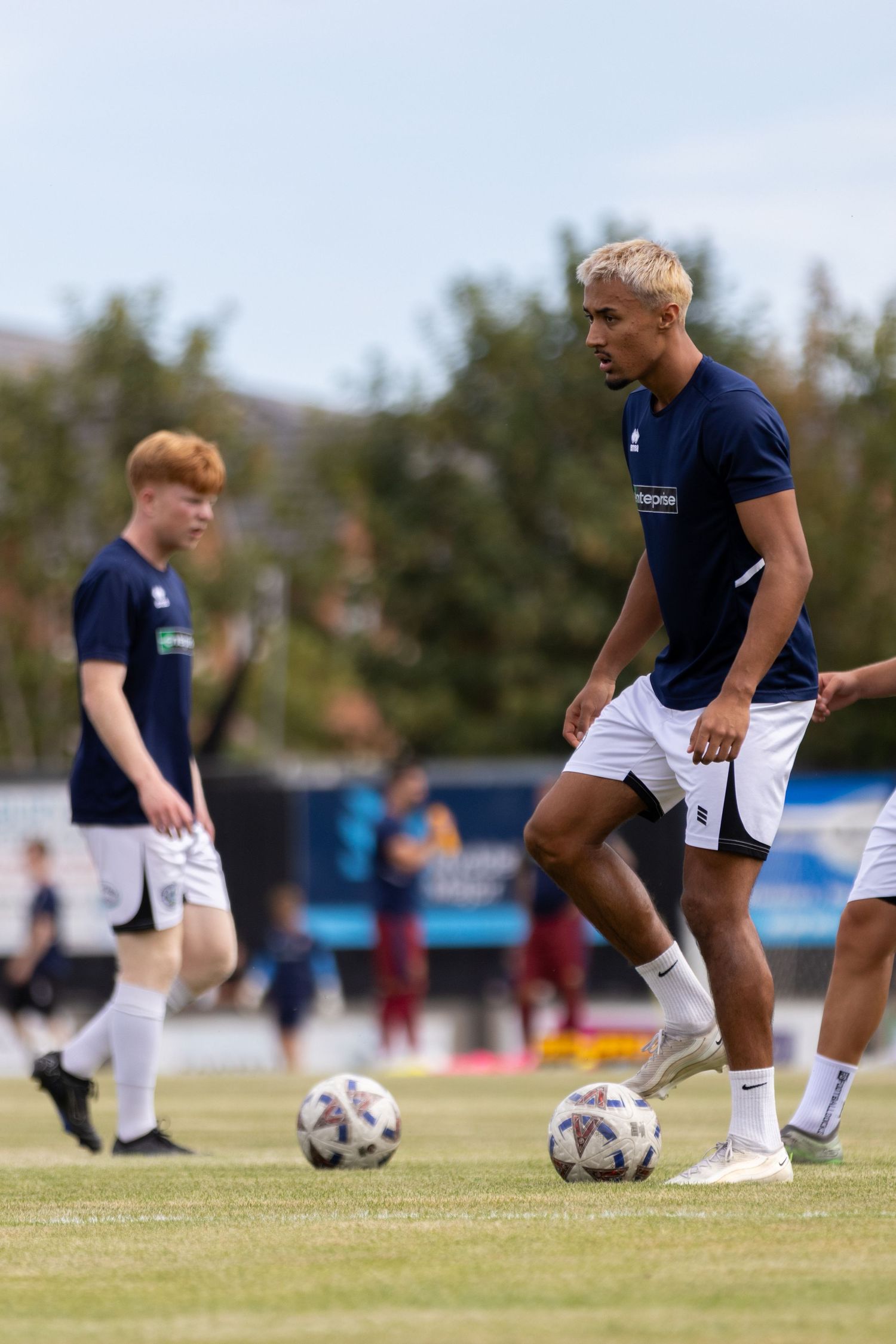 Two soccer players in navy blue uniforms practice dribbling footballs on a green pitch during a sunny outdoor training session.