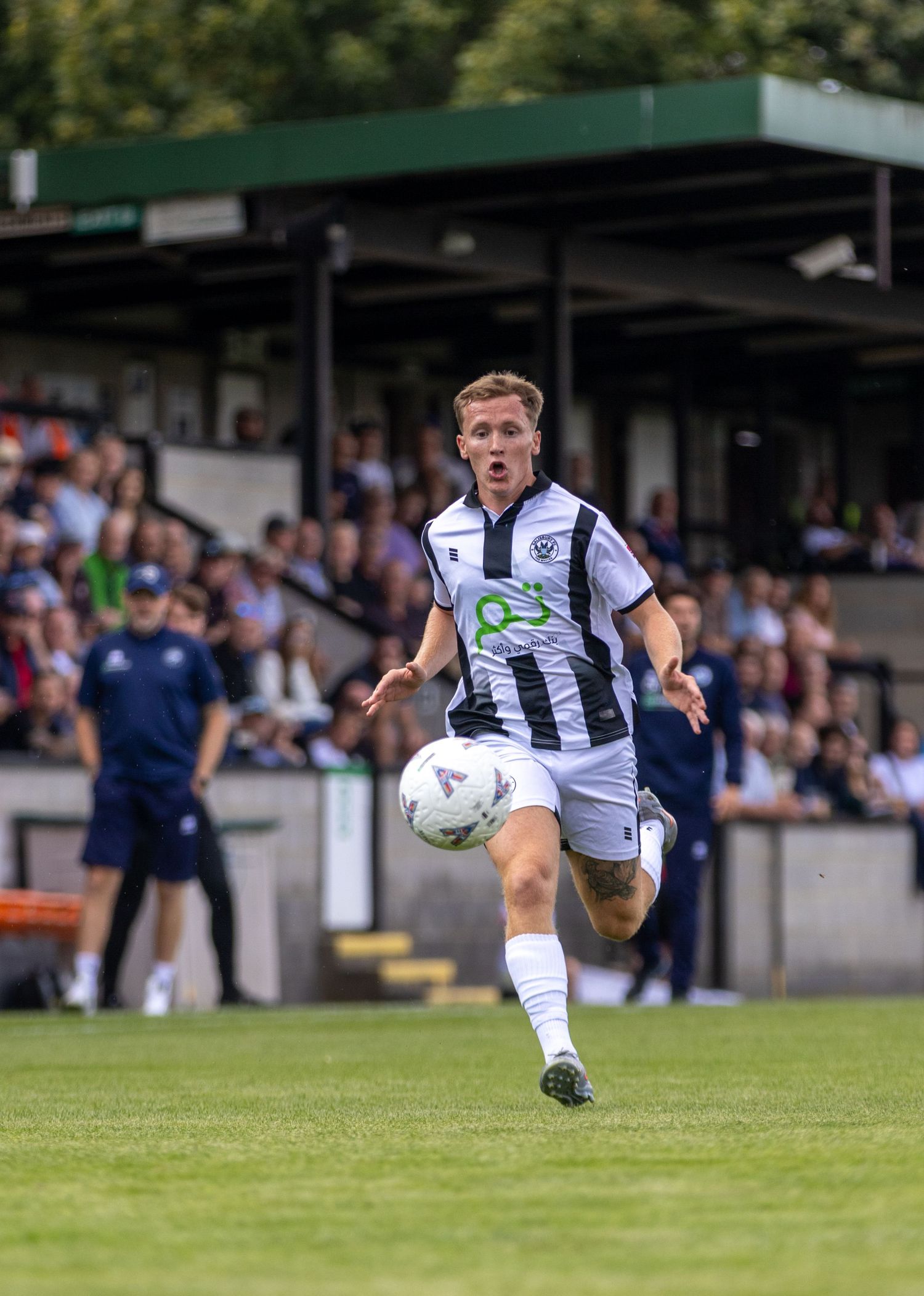 Young footballer in black and white striped kit dribbles the ball forward on a green pitch with crowd in background.