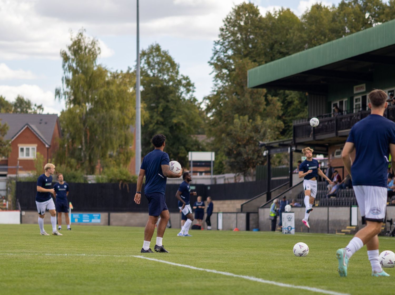 Soccer players in navy blue uniforms warming up on a green pitch before a match at a small stadium.