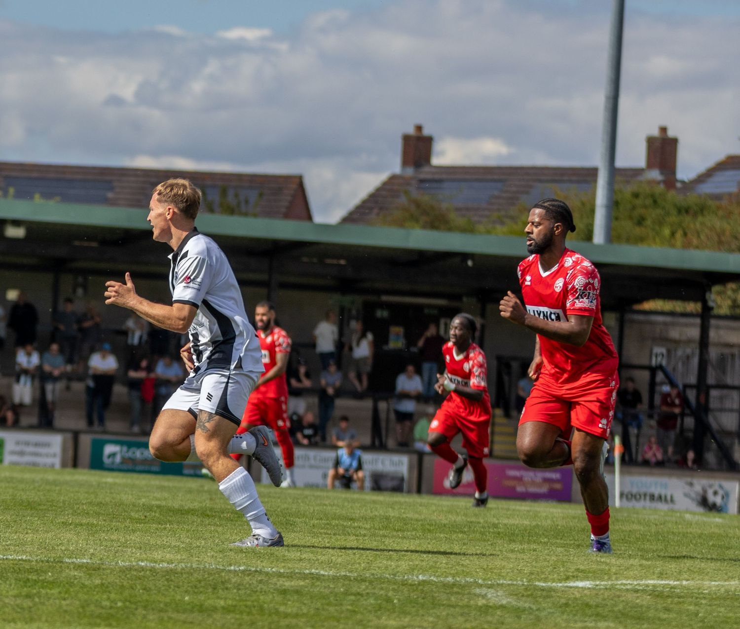 Footballer in white kit celebrates while opponent in red chases during a sunny football match.