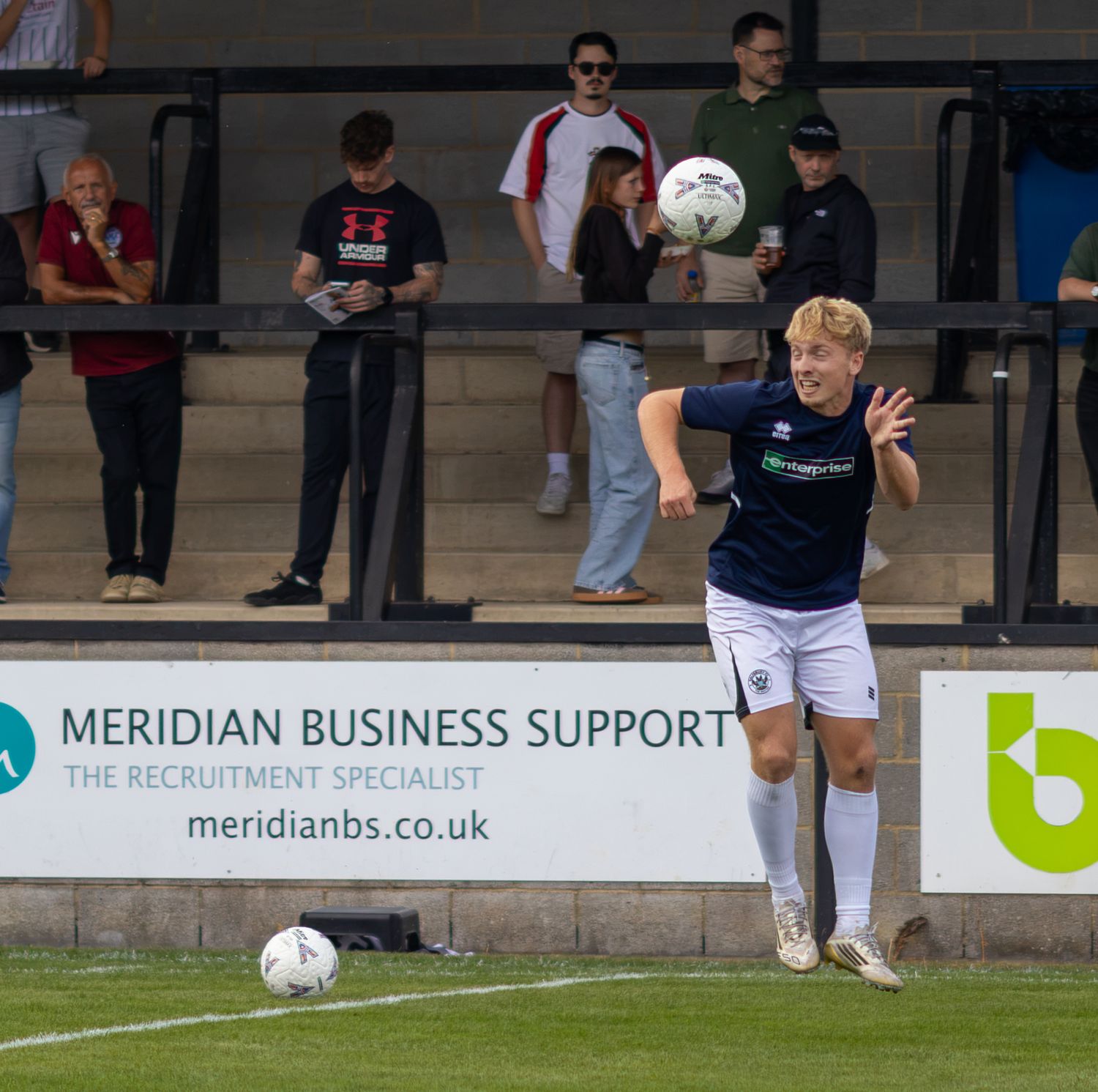 A footballer in white kit heads the ball during a match near a Meridian Business Support advertisement board.