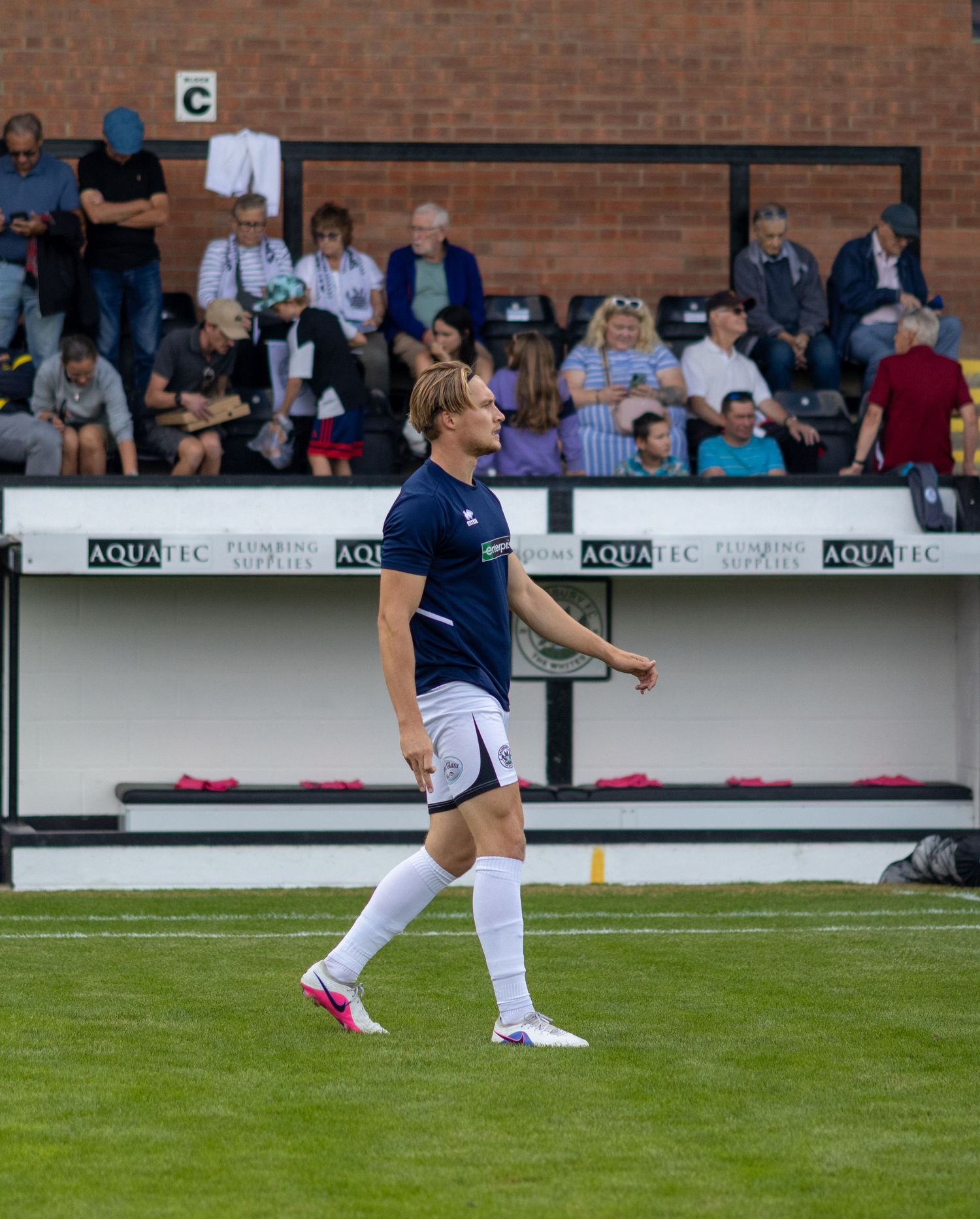 A footballer in navy blue kit and white socks walks across a green pitch during a match with spectators in the background.